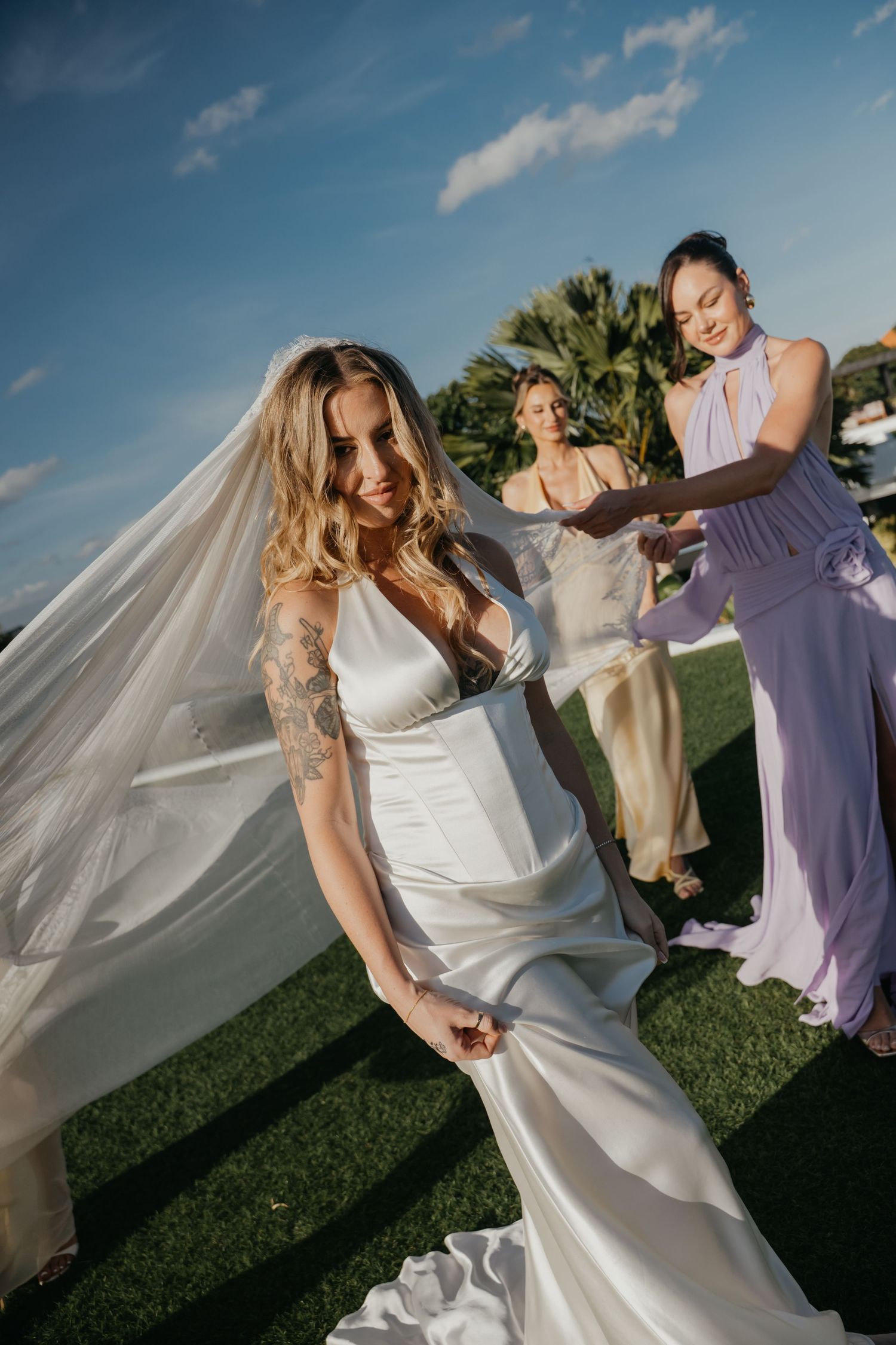 Bride in white satin gown with veil being adjusted by bridesmaids in lavender and gold dresses.