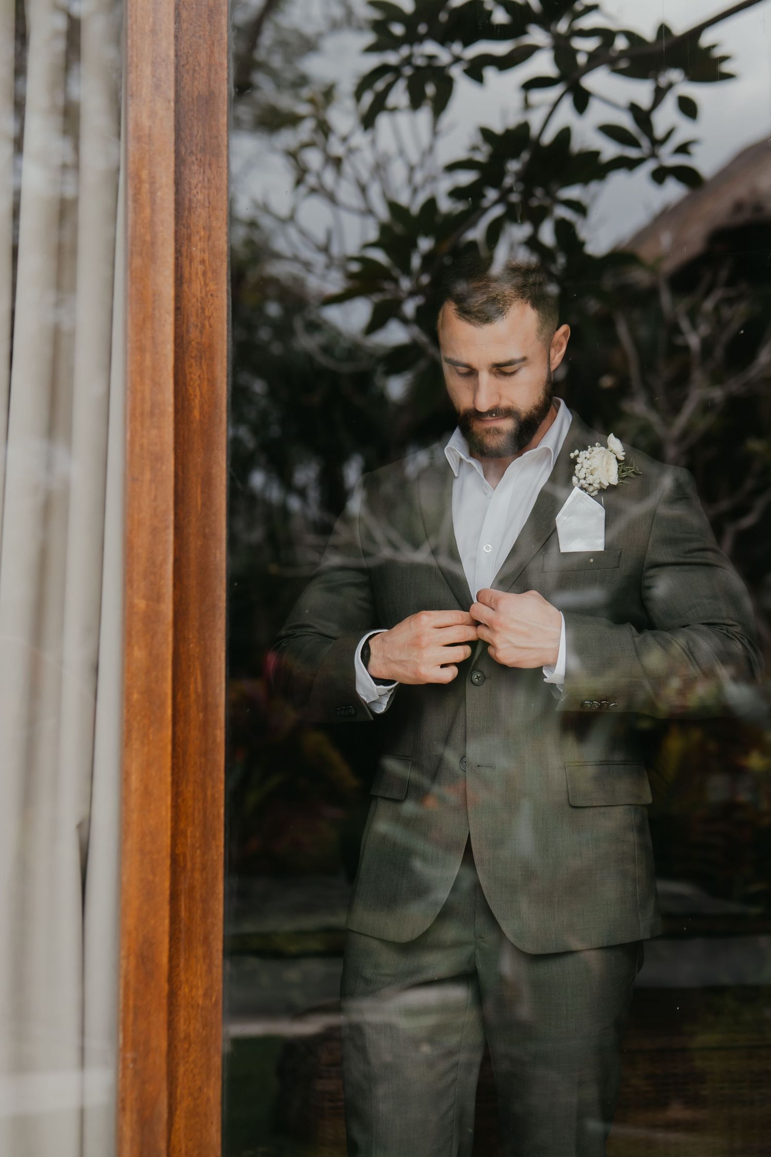 Dapper bearded groom in gray suit with boutonniere buttons his jacket beside a wooden framed glass door.