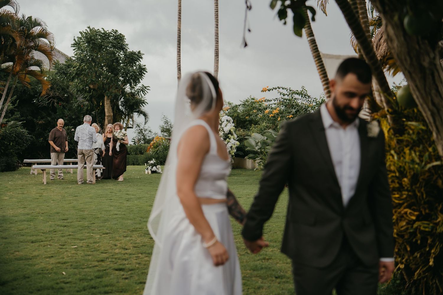 Newlywed couple holds hands walking away from floral arch as wedding guests gather on garden lawn.