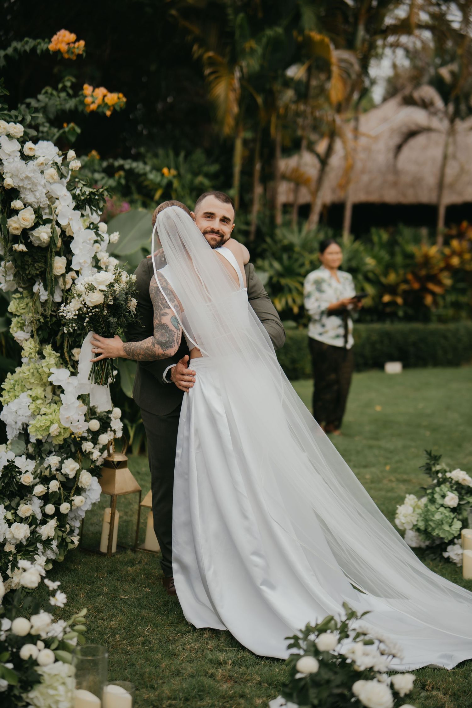 Smiling groom with tattoos embracing bride in elegant white gown and veil at outdoor floral ceremony.