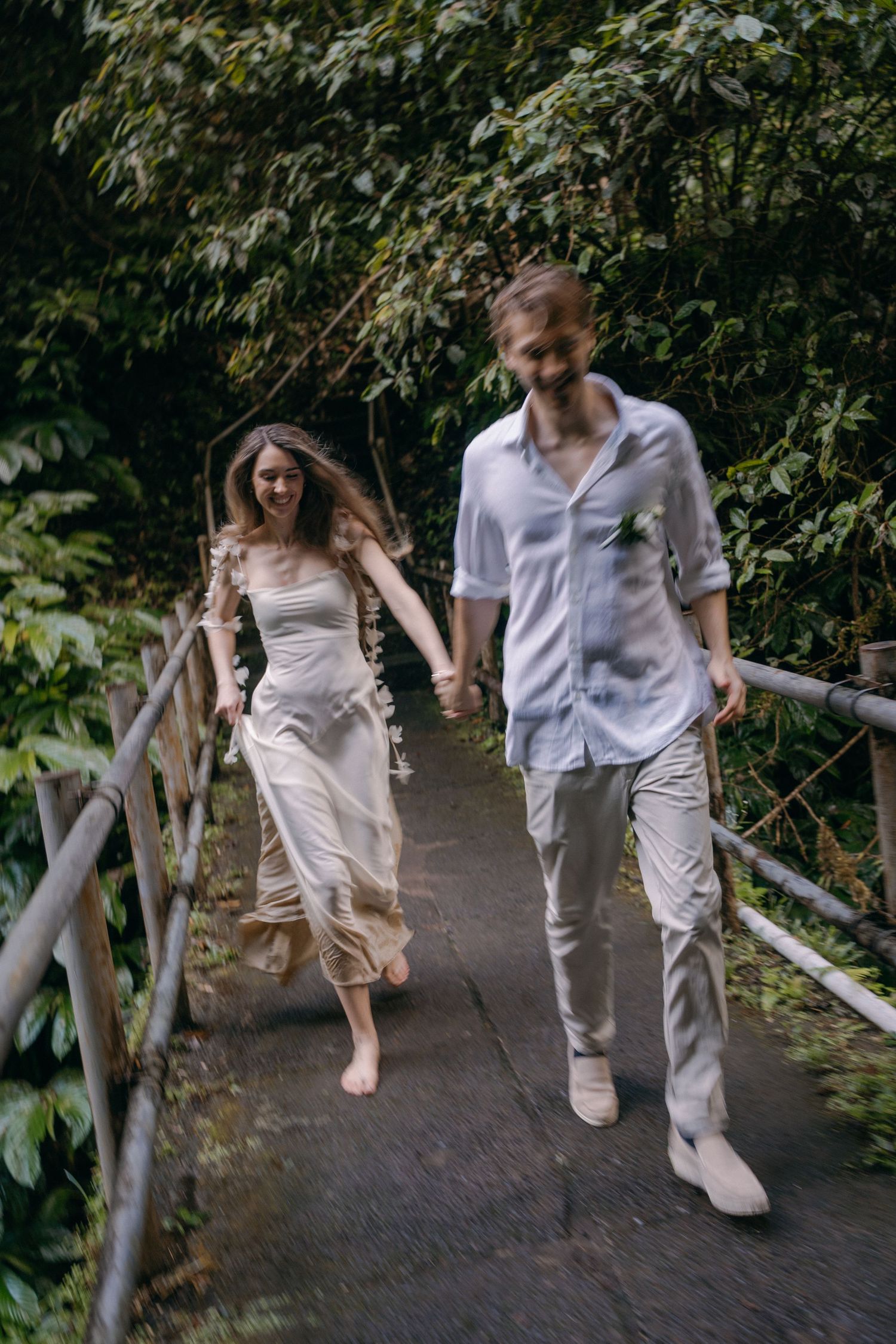 A couple in light summer attire walking barefoot and holding hands on a forest path surrounded by green foliage.