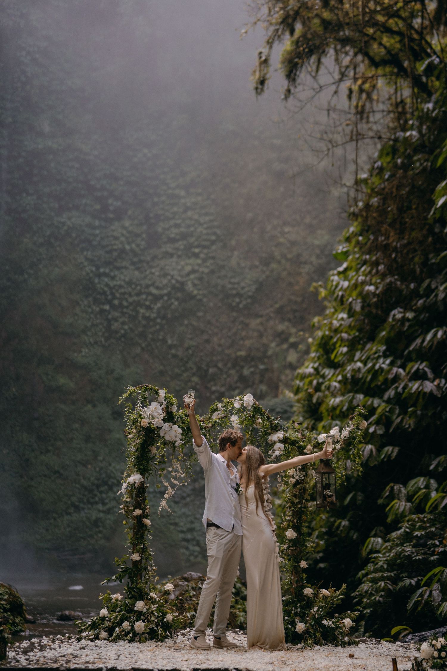 Couple celebrates under floral arch in misty forest setting with sunlight streaming through trees and lush greenery.
