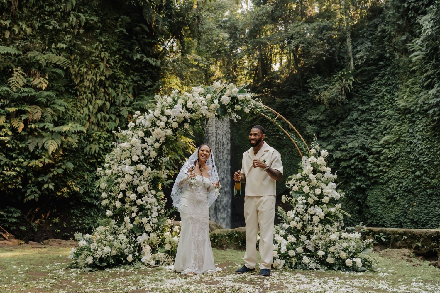 Happy newlyweds celebrating under a white floral arch surrounded by lush tropical greenery in Bali.