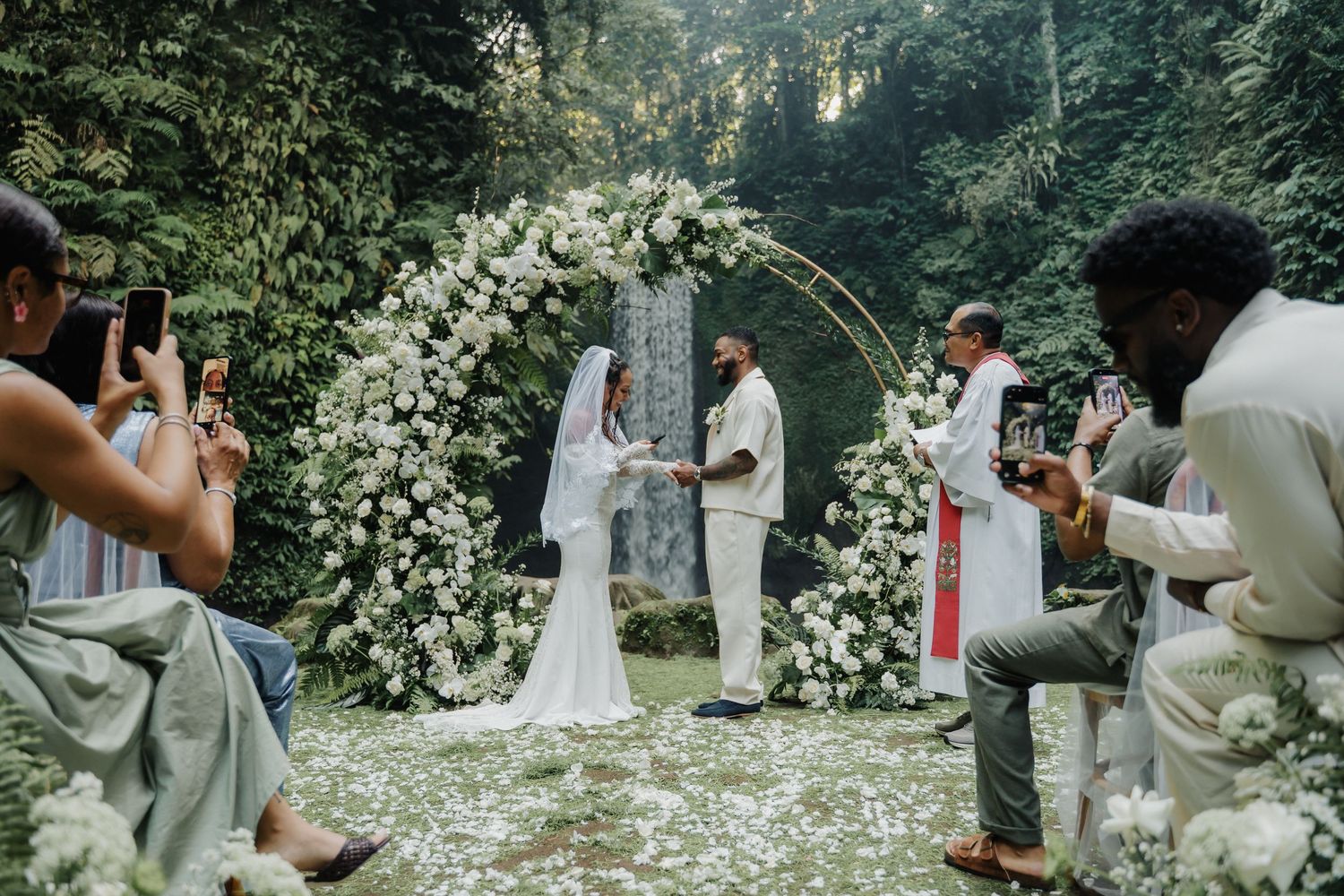 Bride and groom exchange vows under a circular floral arch with waterfall backdrop in tropical forest.