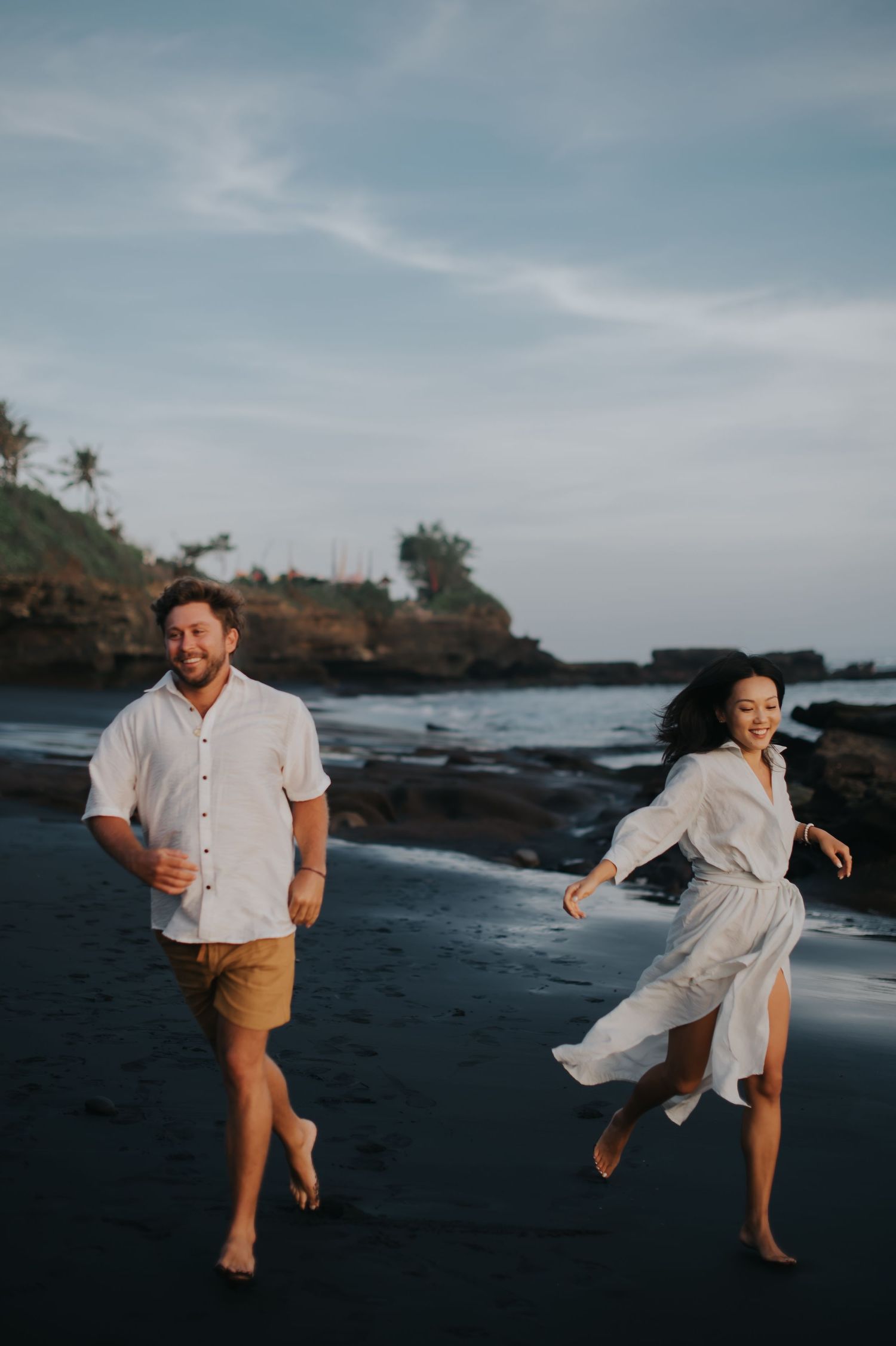 Two people dance and laugh together on a dramatic volcanic beach shoreline at sunset.