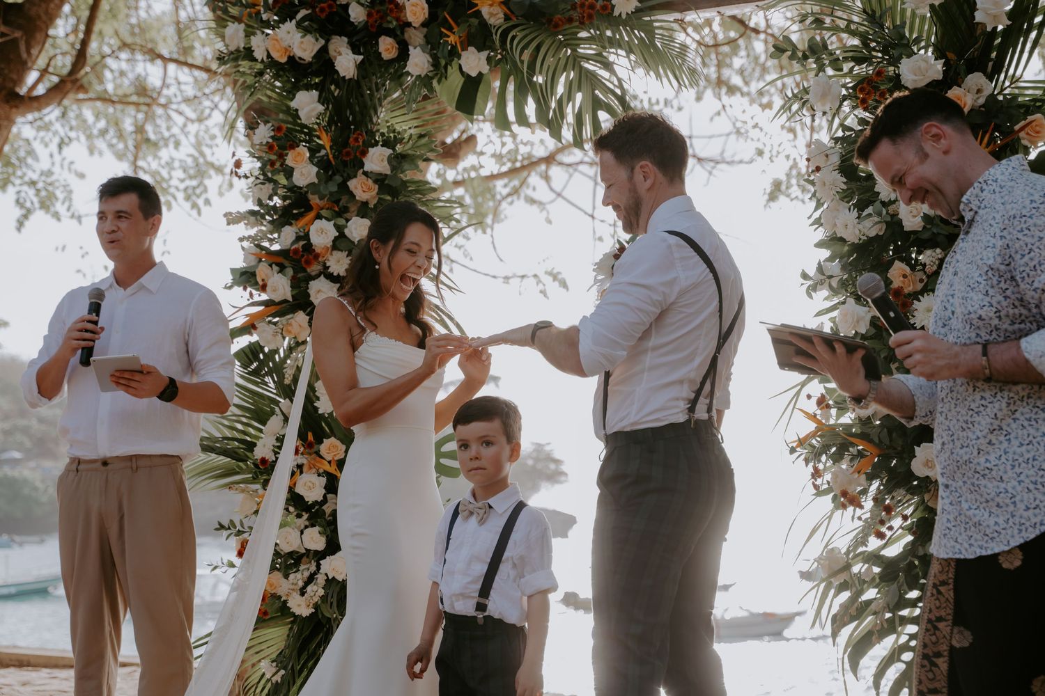 A beach wedding ceremony takes place under a floral arch with tropical decor and ocean views in the background.