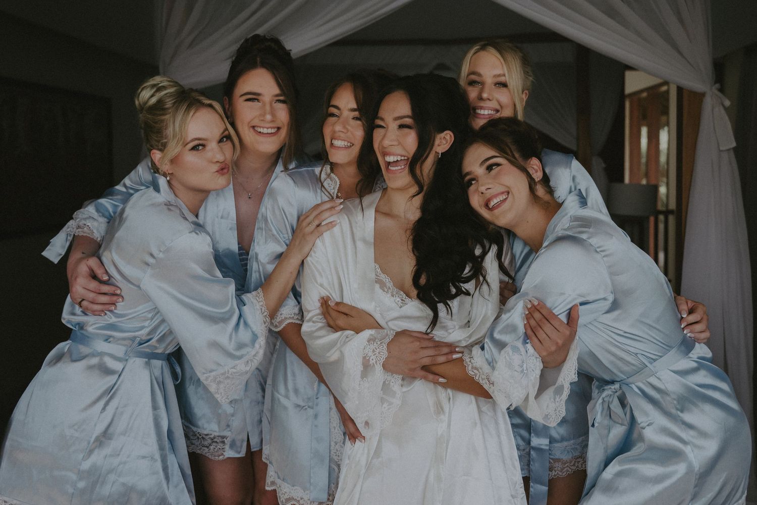 Bridesmaids in matching silk robes surround bride in white robe while laughing and hugging during wedding preparations.