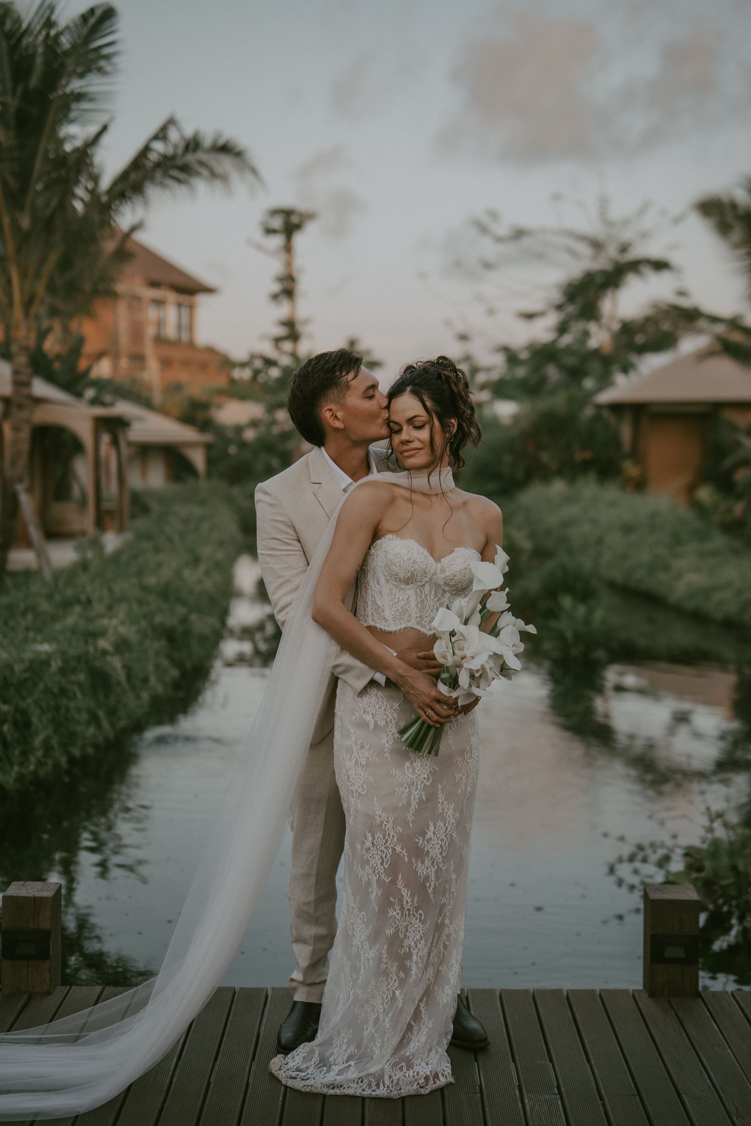 A romantic wedding couple shares intimate moments on a wooden dock by a tropical resort at sunset.