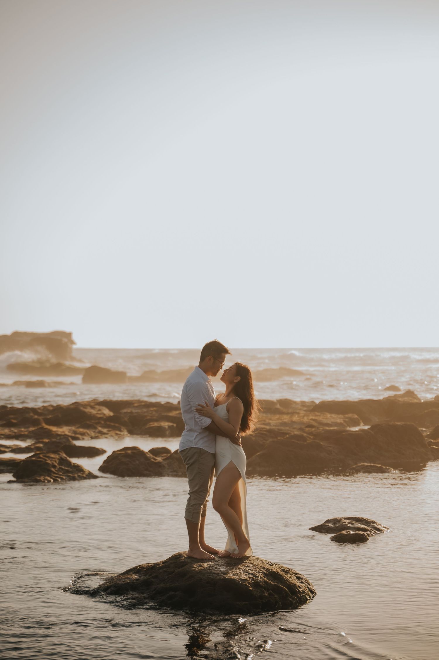 Romantic couple embracing on a rock at the beach during golden hour sunset with ocean waves in the background.
