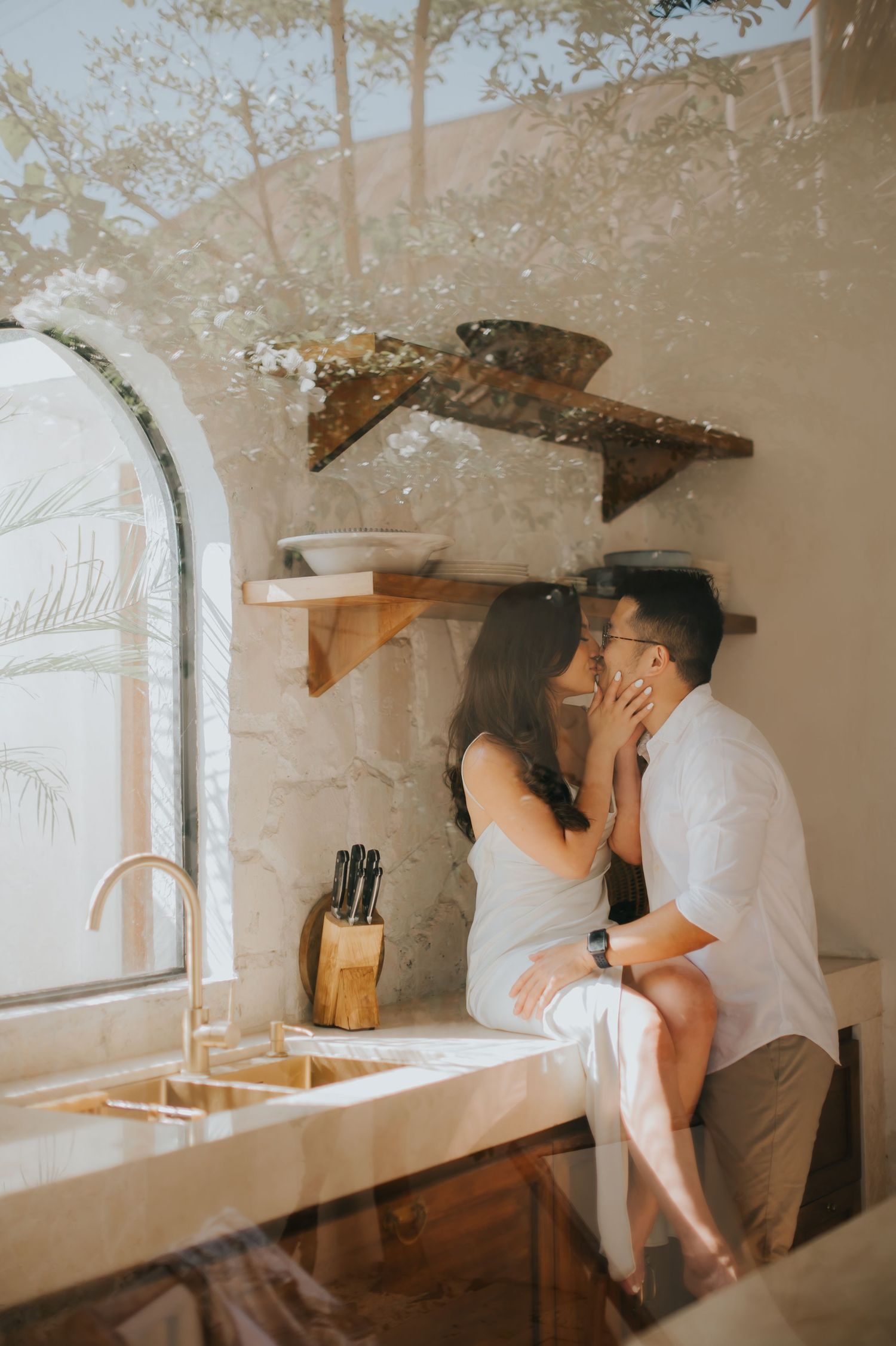 Couple sharing a romantic kiss in a rustic kitchen, sitting on the counter near an arched window with warm natural light.