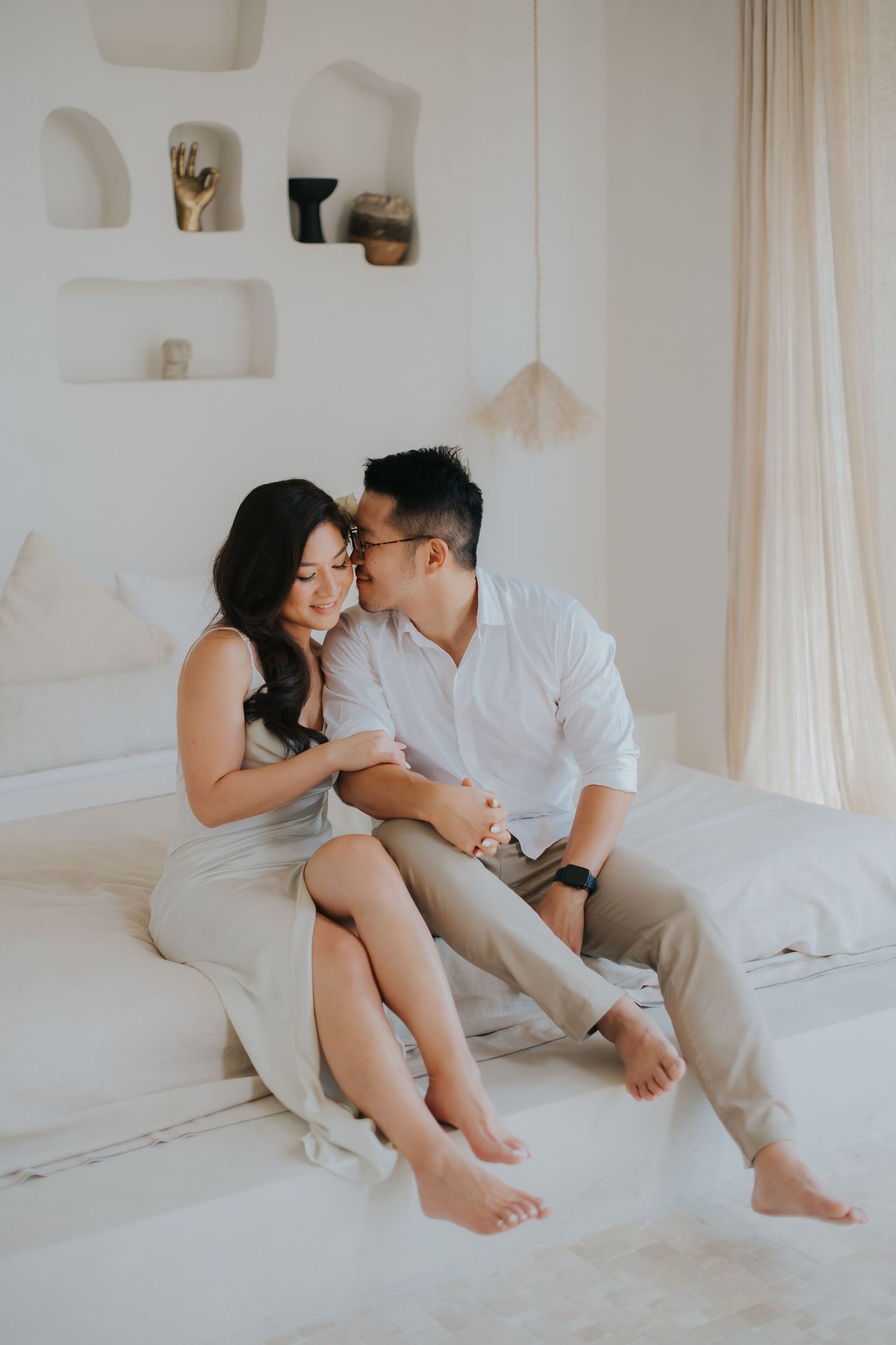 Couple sharing a tender moment sitting on a white bed in a bright, minimalist bedroom with neutral tones.
