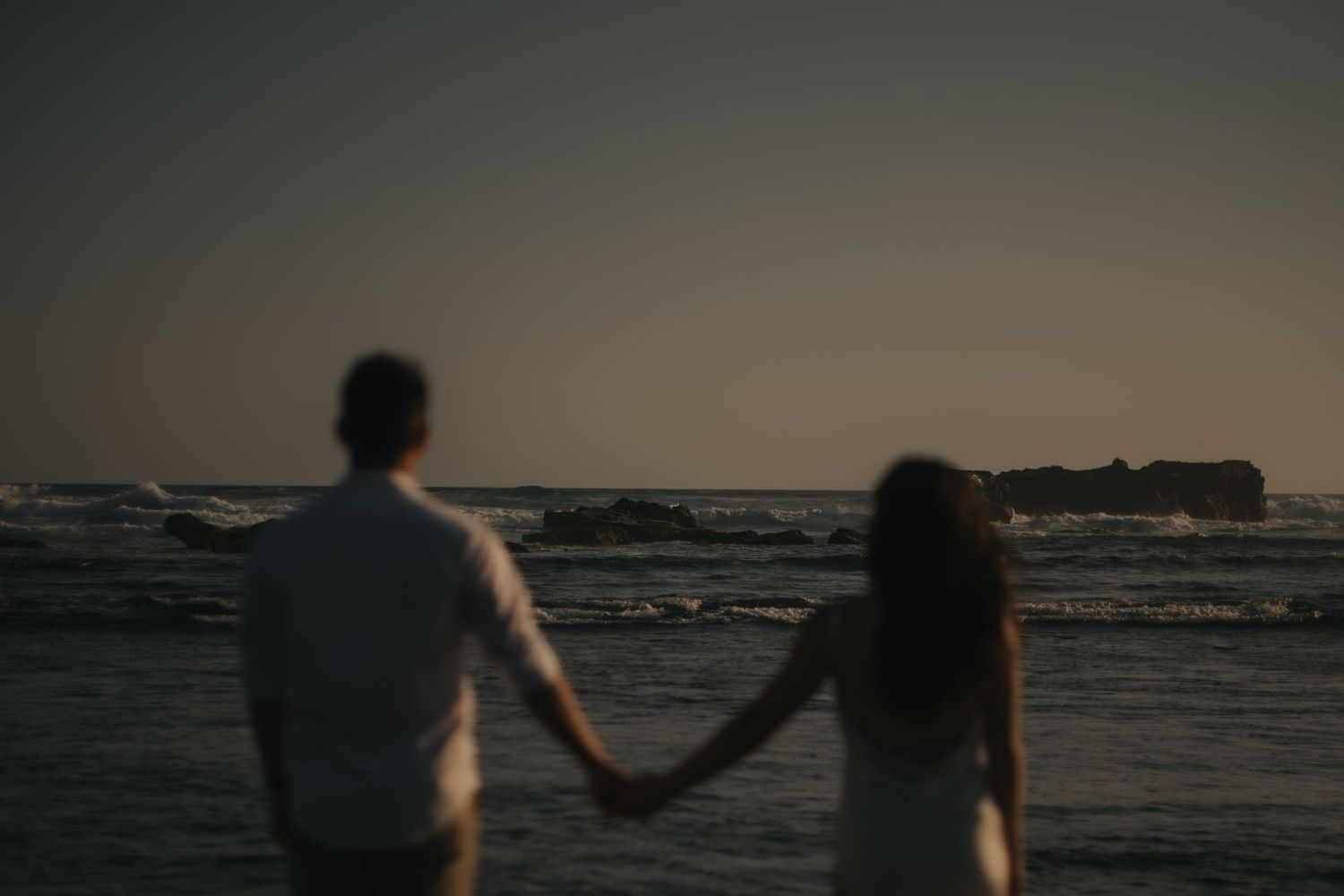Couple holding hands on a rocky beach at dusk, gazing at the ocean horizon in a romantic silhouette.