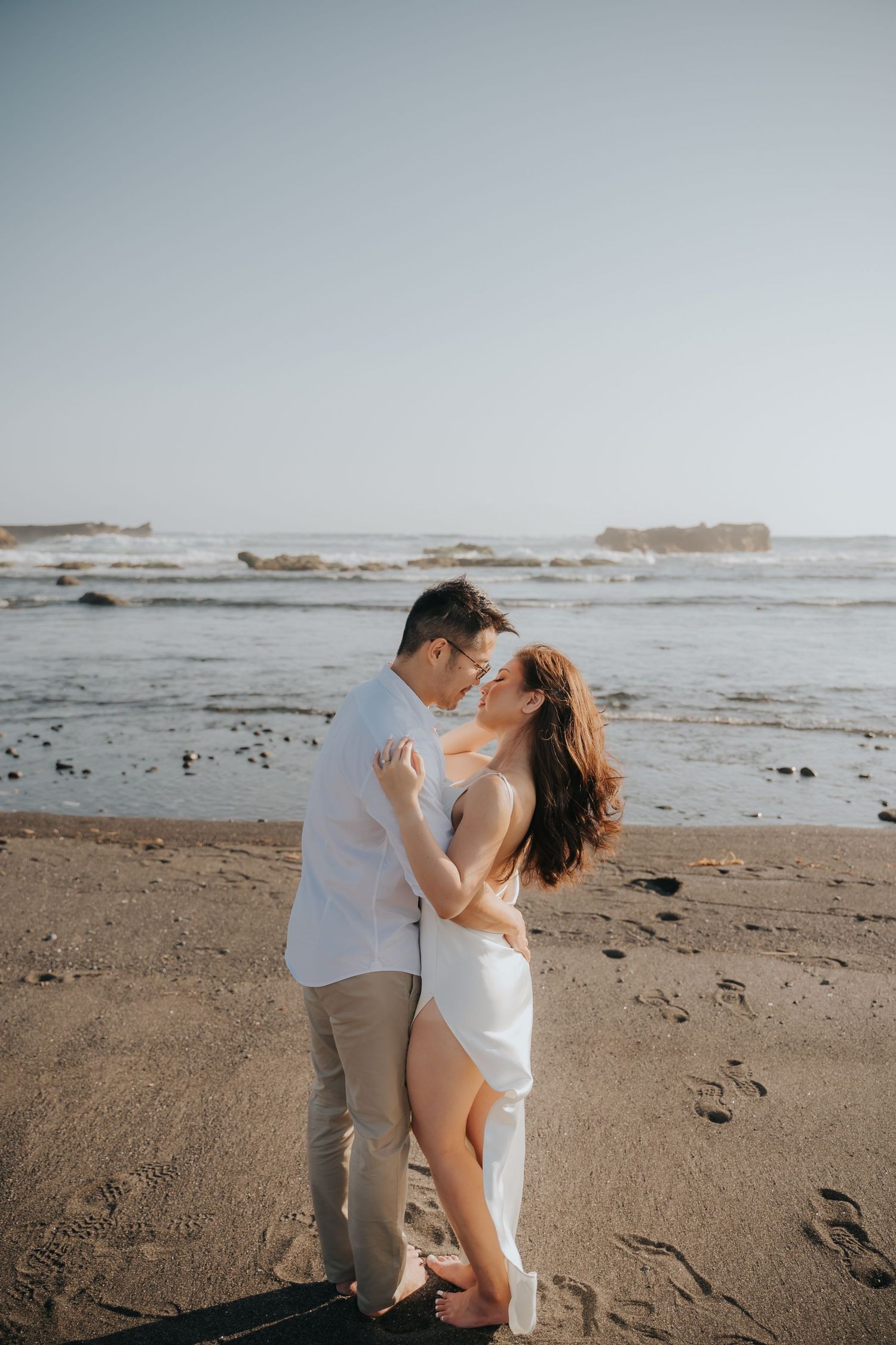 Romantic couple embracing and kissing on a sandy beach with ocean waves and rocks in the background.