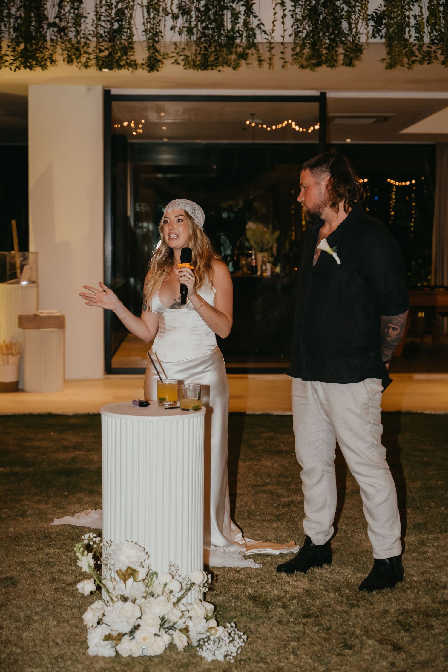 Bride in white satin dress speaks into microphone beside groom at outdoor wedding reception.