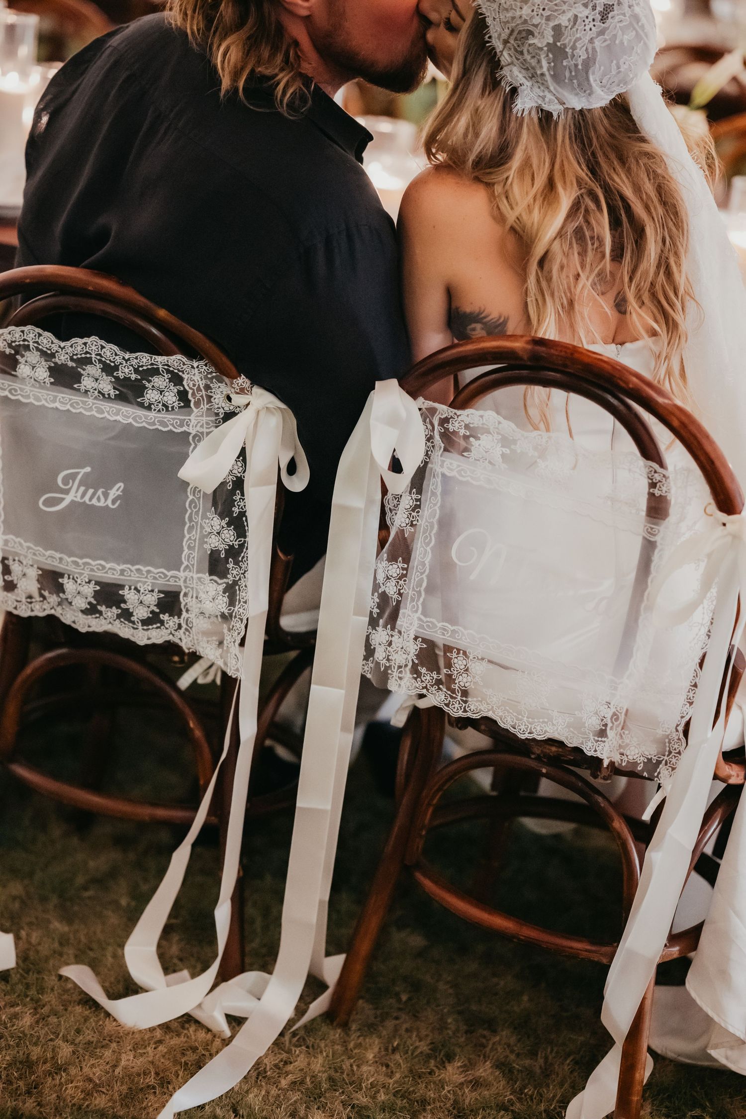 Bride and groom share a kiss behind lace-decorated Just Married chairs at wedding reception.
