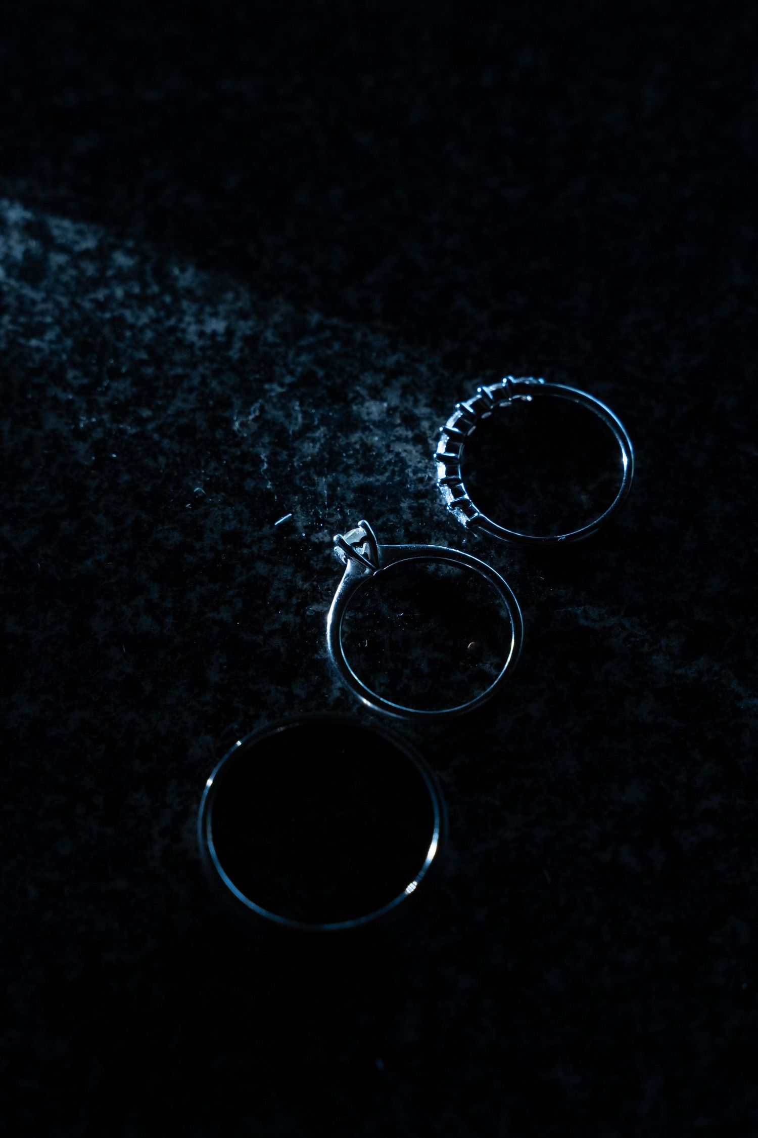 Three silver wedding rings with diamonds displayed on dark stone surface in dramatic lighting.