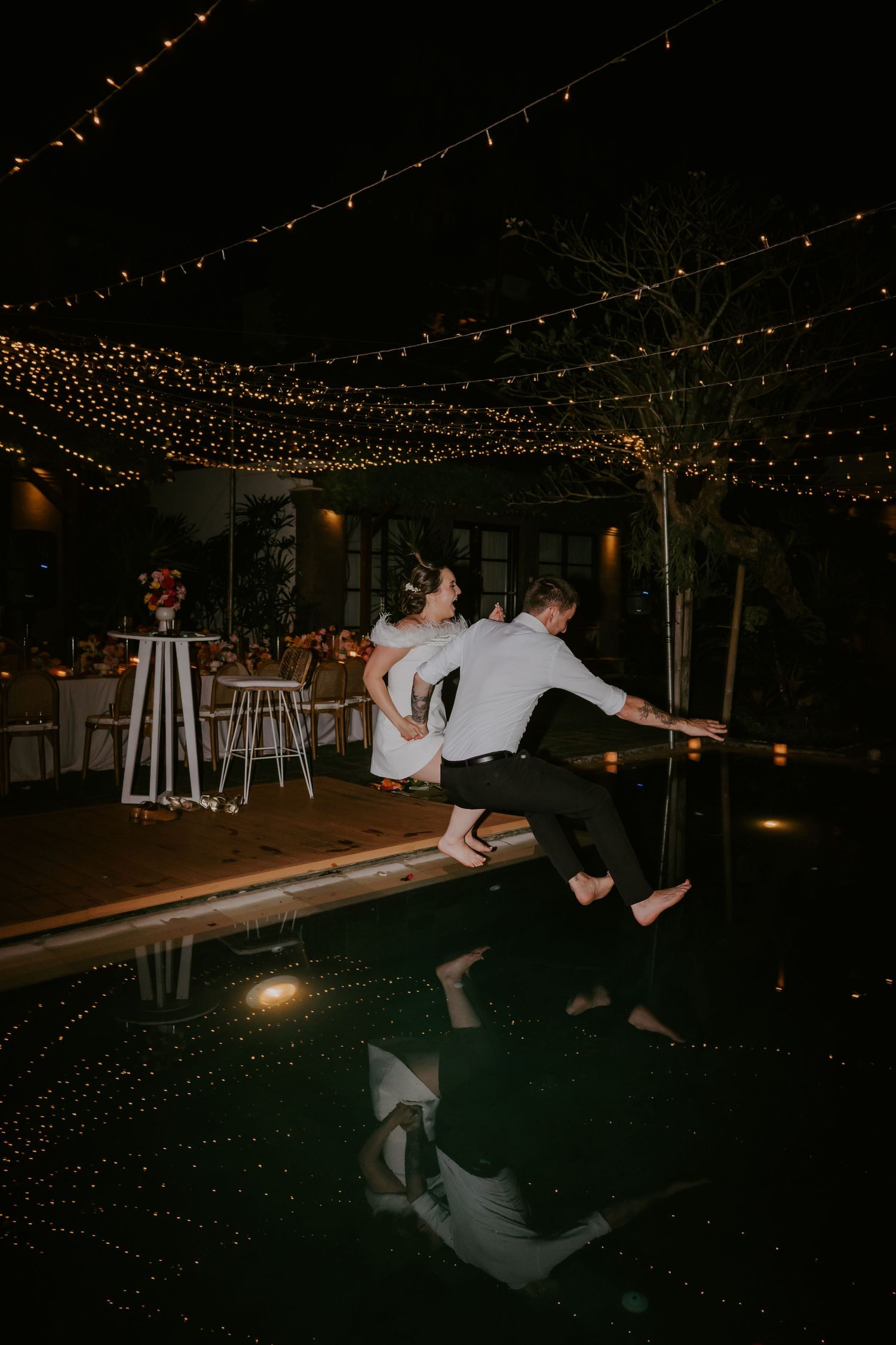 Bride and groom jumping into a pool at night under twinkling string lights at their wedding reception.
