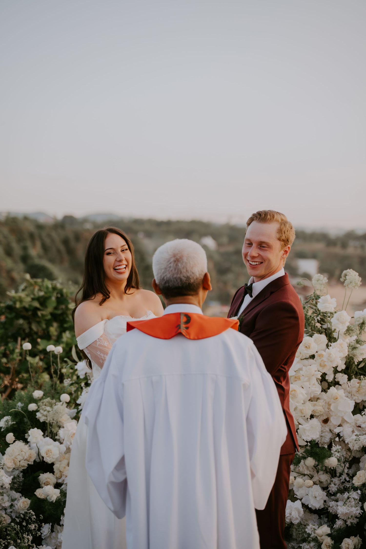Smiling bride and groom exchange vows with officiant at outdoor wedding ceremony with white flowers.