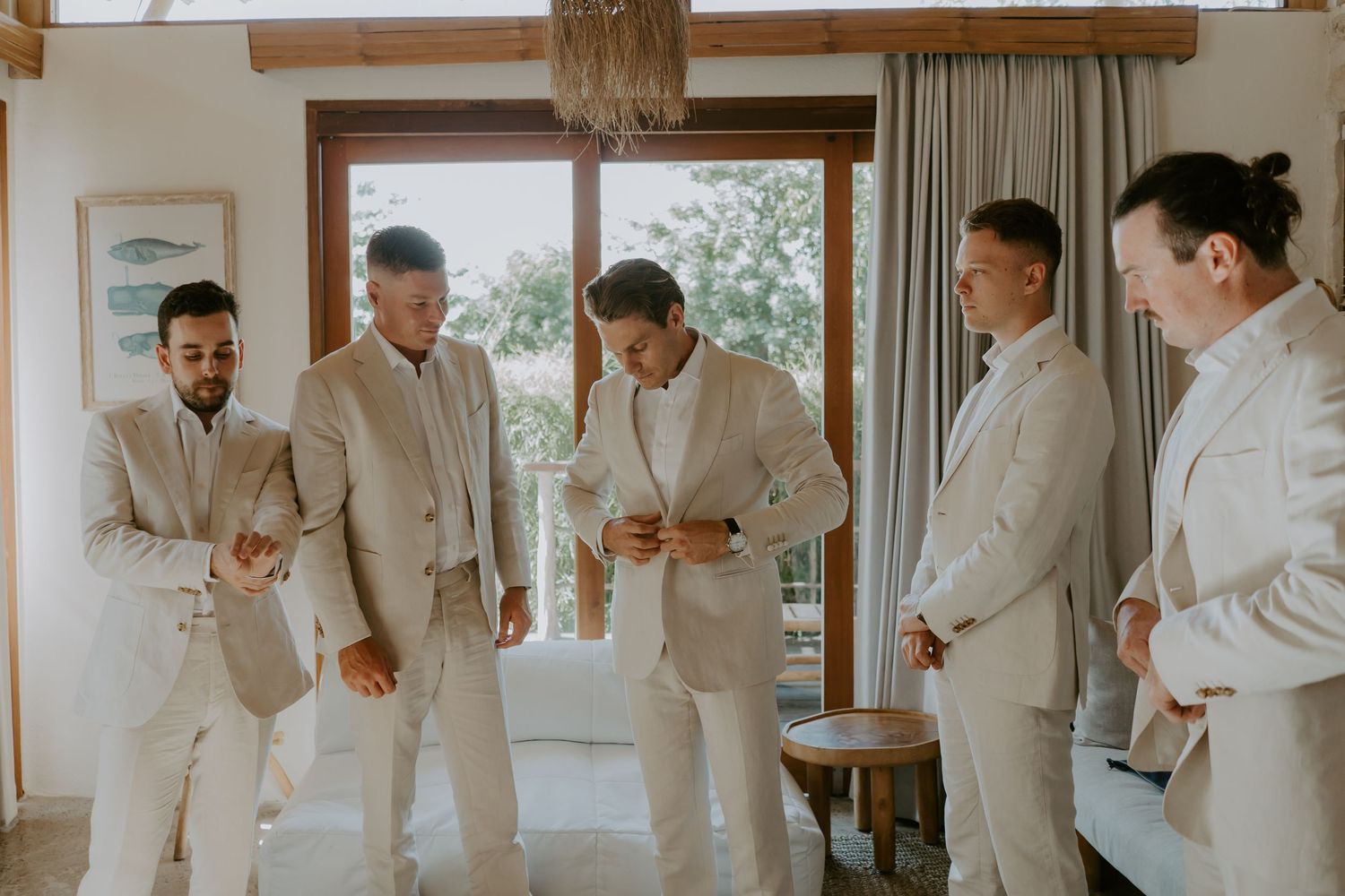 Five groomsmen in matching beige suits prepare together in bright room before wedding ceremony.