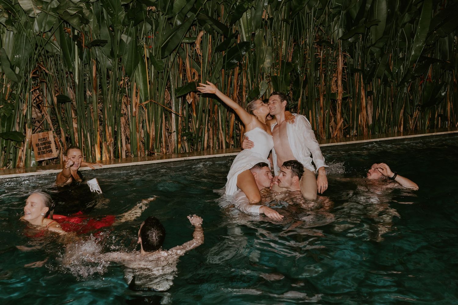 Bride and groom celebrate in pool surrounded by joyful guests at tropical wedding after-party.