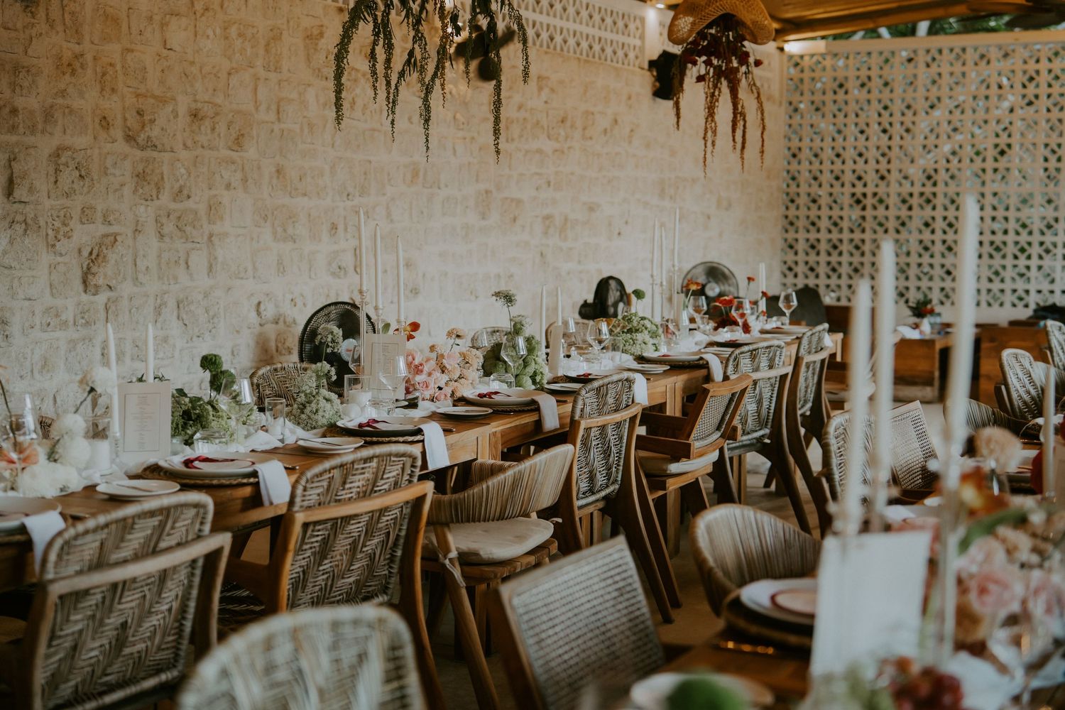 Elegant wedding reception tables with floral centerpieces, candles, and wicker chairs in a stone-walled venue.