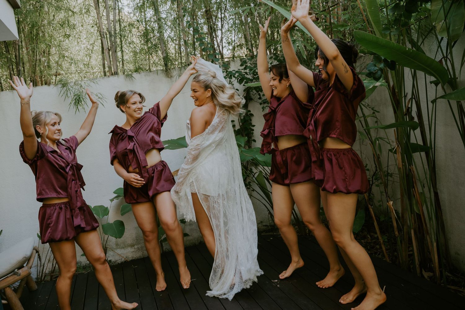 Bride and bridesmaids in burgundy robes celebrate together outdoors surrounded by tropical greenery.