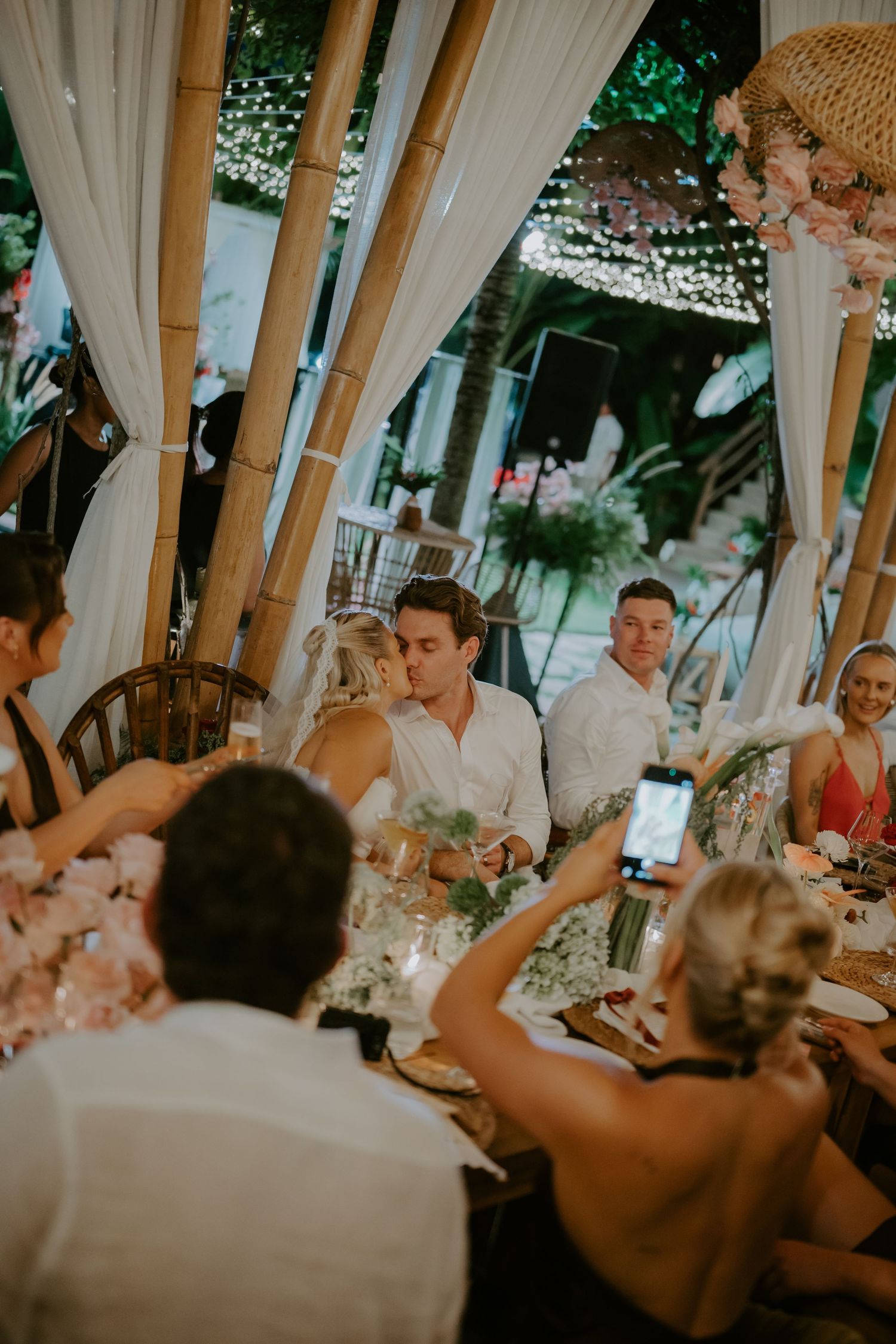 Bride and groom share a kiss at decorated reception table while guests capture the moment.