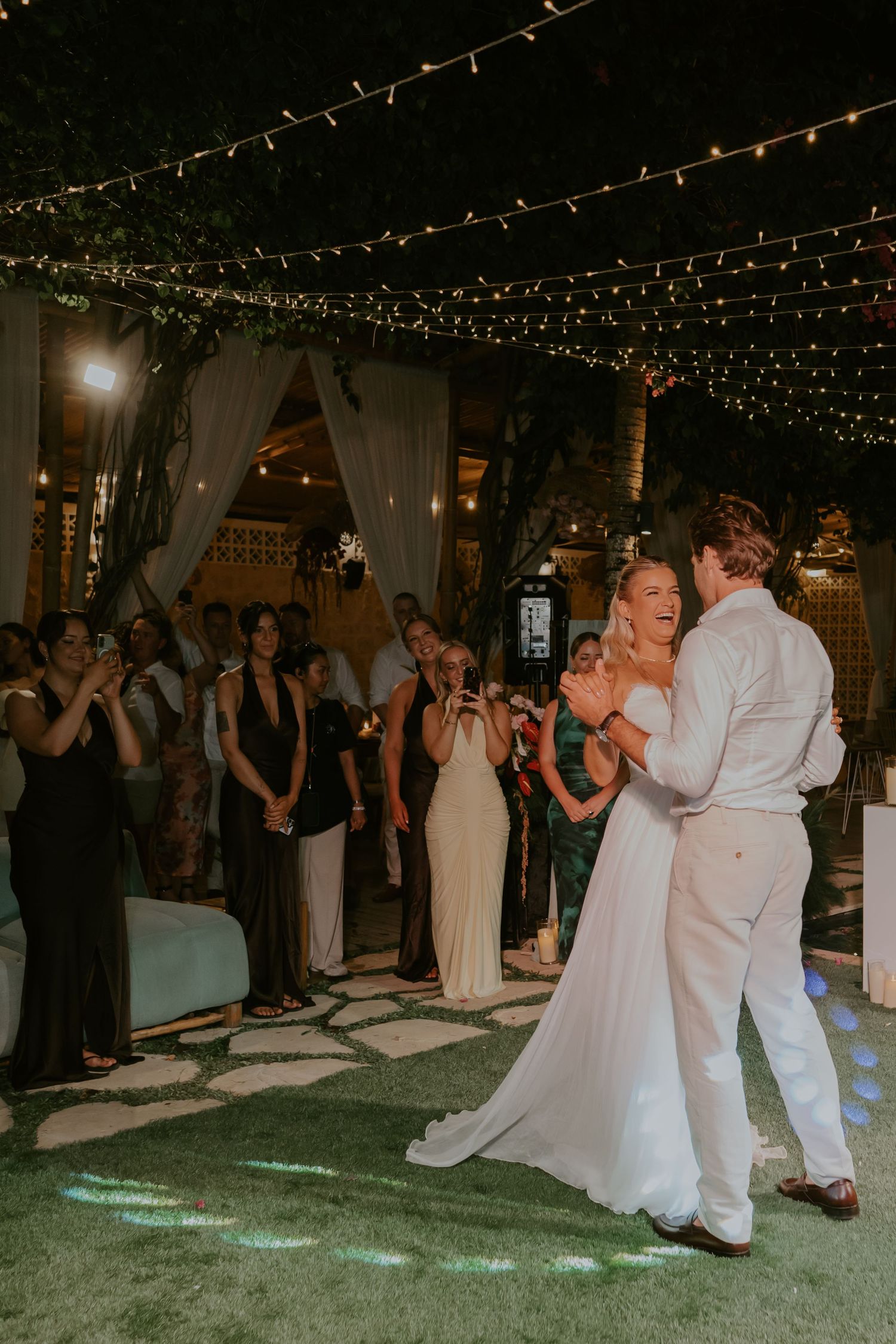 Bride in white gown and groom dance their first dance on outdoor dance floor under string lights.