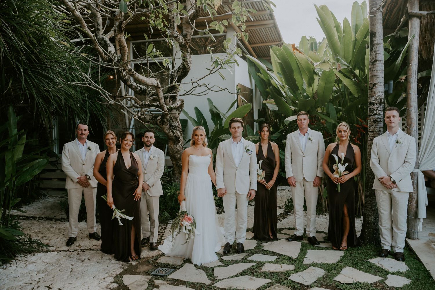 Wedding party poses on stone path surrounded by tropical gardens at outdoor venue.