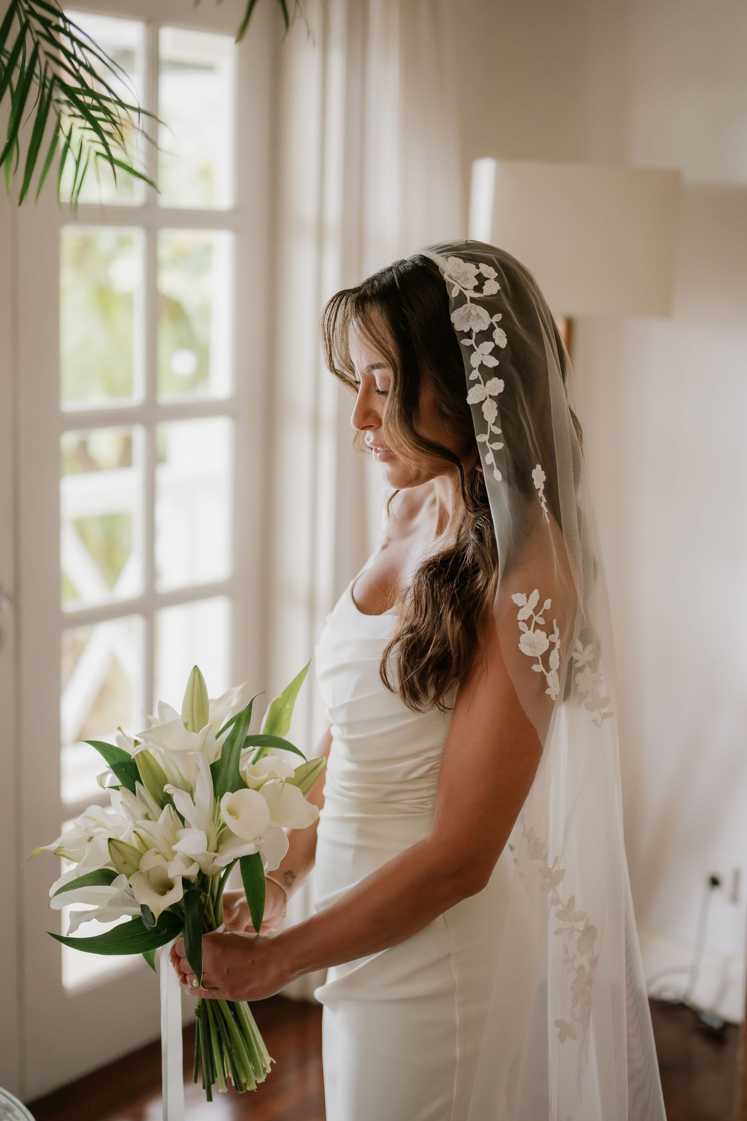 Bride in white gown and floral veil holding white lily bouquet by window.