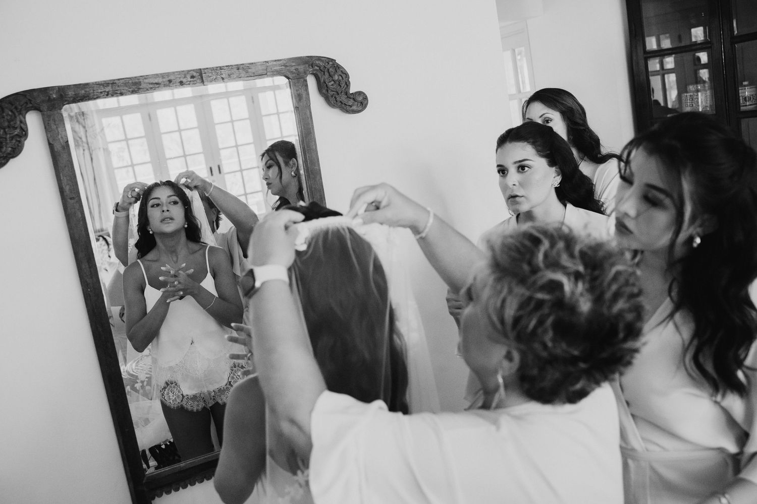Bridesmaids help bride put on veil in front of mirror in black and white wedding preparation photo.