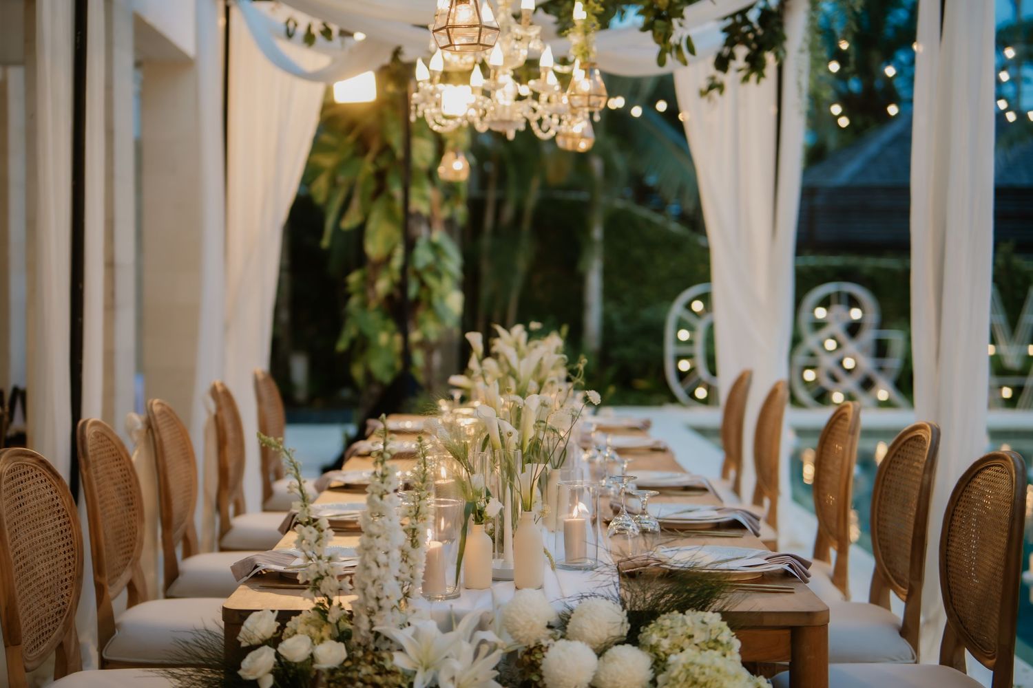 Elegant wedding reception table with white floral centerpiece and chandelier under draped canopy.