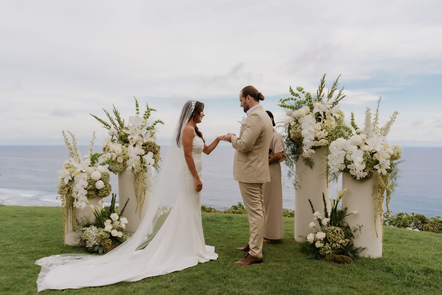 Bride and groom exchanging vows at a clifftop ceremony with white floral arrangements and ocean backdrop.