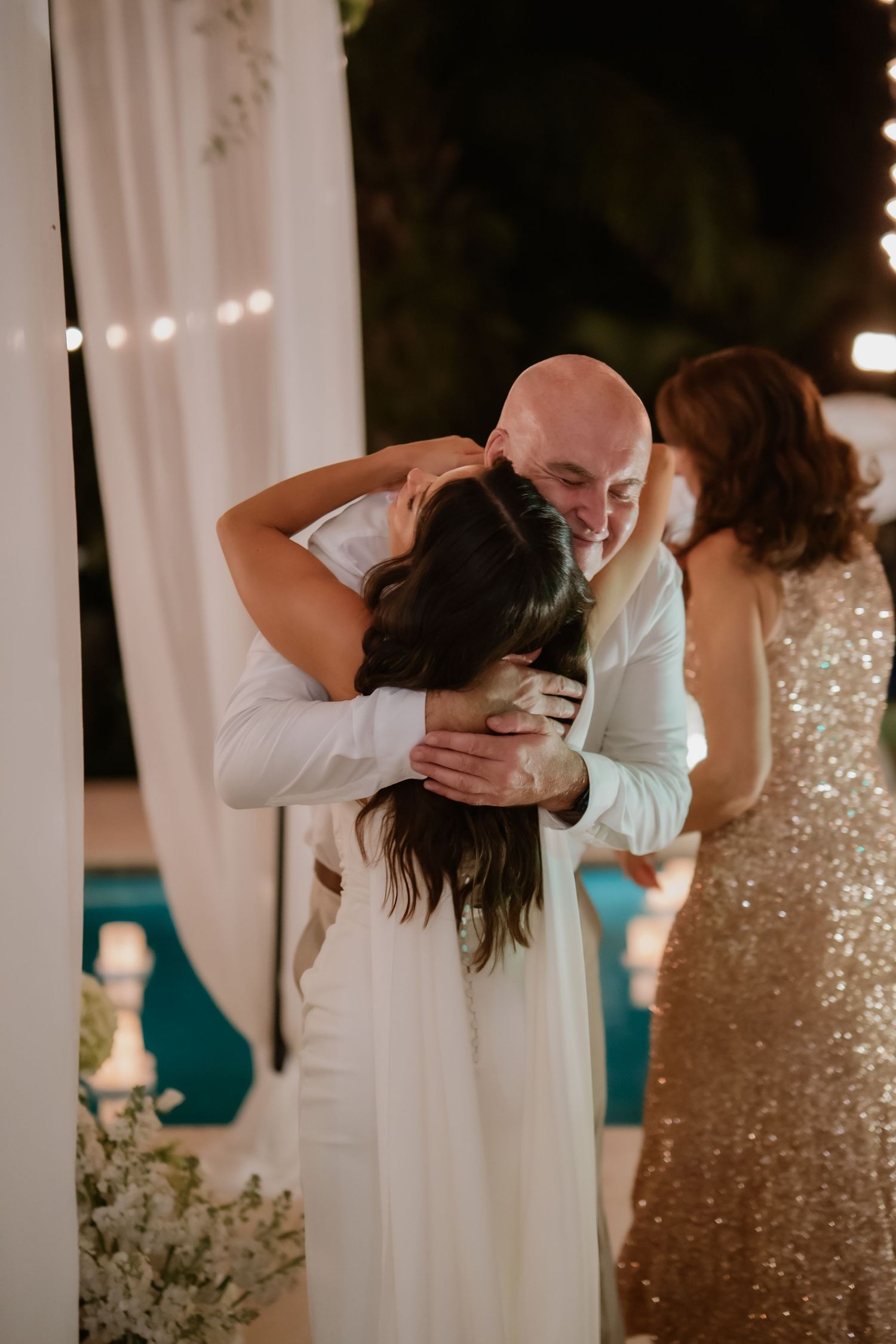Bald groom embracing bride in white dress under floral arch at romantic nighttime wedding ceremony.