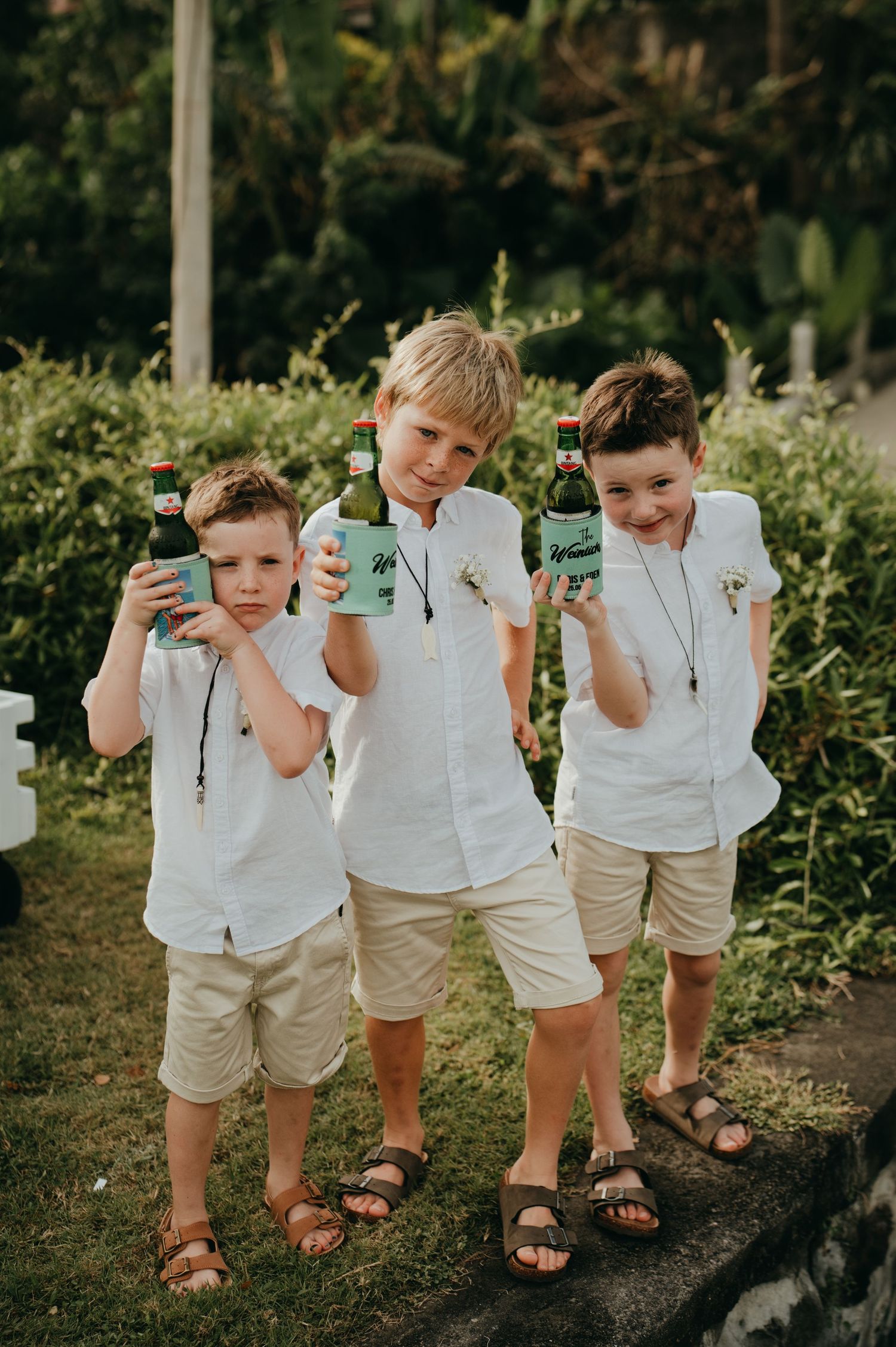 Three young boys in white shirts and tan shorts holding green beer bottles at an outdoor wedding celebration.