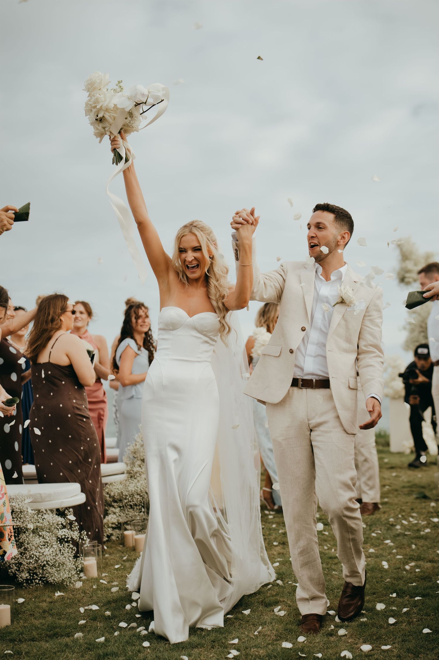 Joyful bride raises bouquet while walking with groom through petal toss at outdoor tropical wedding ceremony.