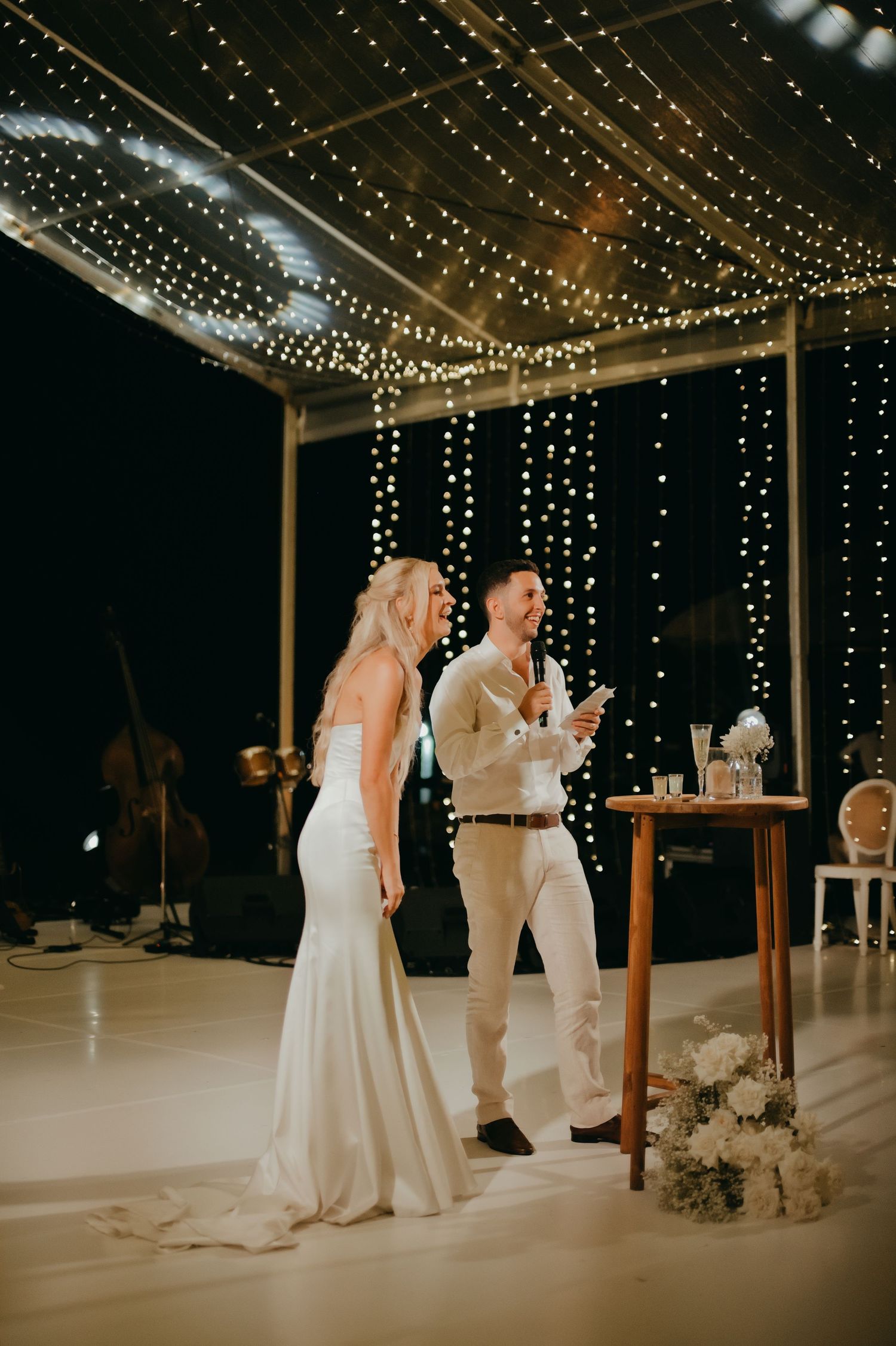Bride and groom laughing together on a dance floor surrounded by twinkling fairy lights at night.