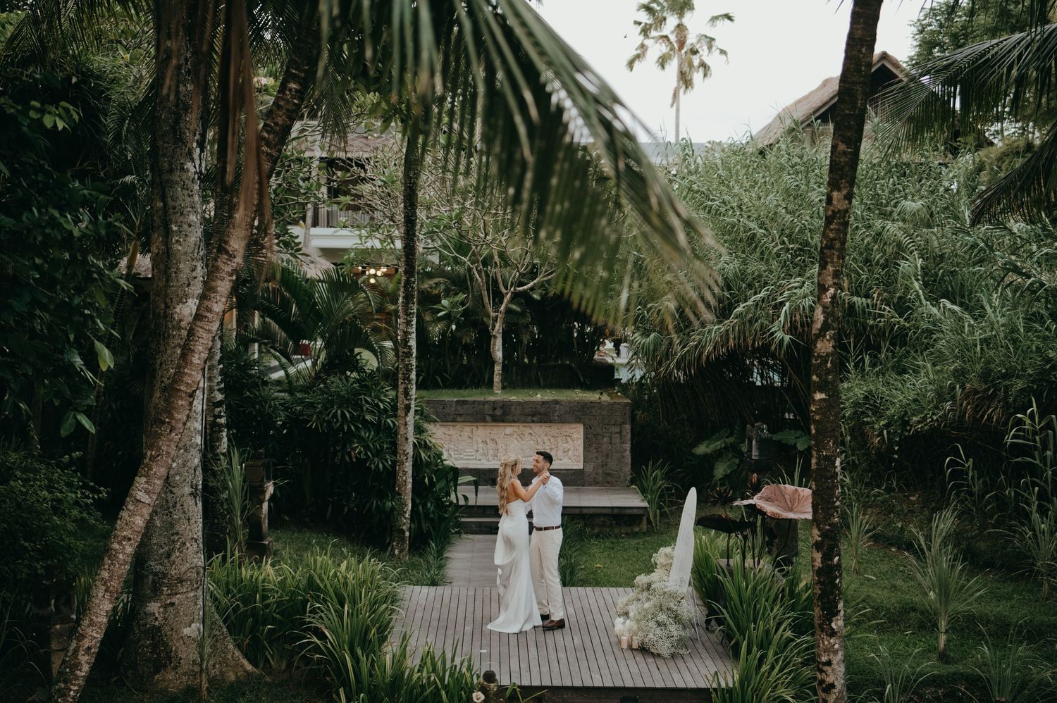 Bride and groom share a tender moment on a wooden deck surrounded by lush tropical garden greenery.