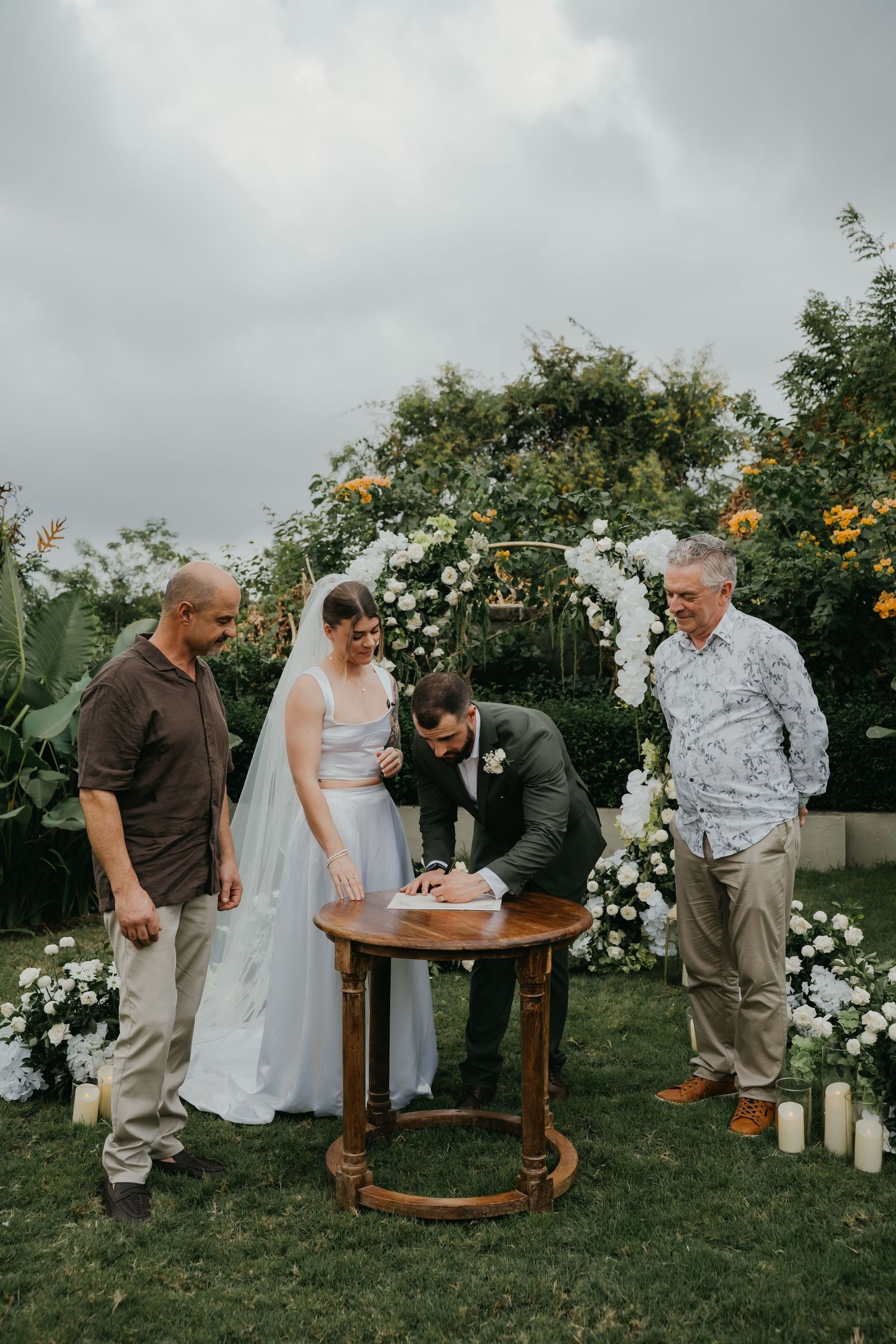 Bride and groom sign wedding register at wooden table flanked by two witnesses under floral arch.