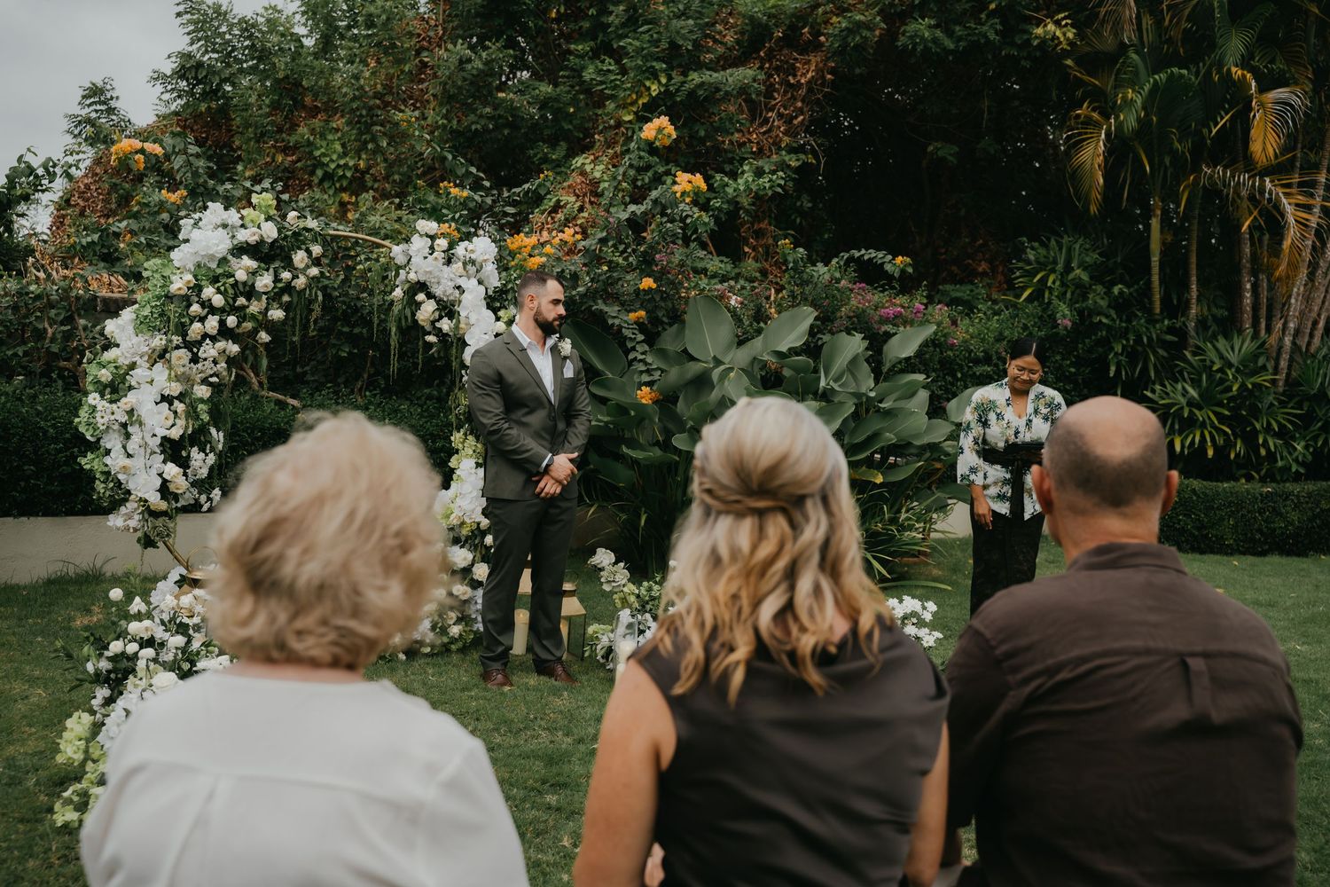 Groom awaits bride at floral arch during intimate outdoor ceremony.