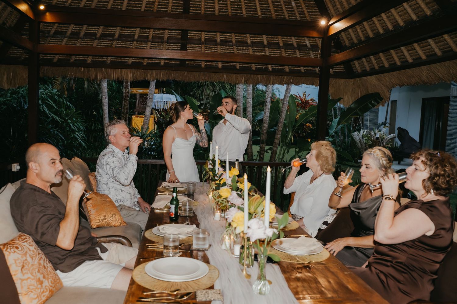 Bride and groom celebrate with small group of guests at candlelit outdoor Bali reception dinner.