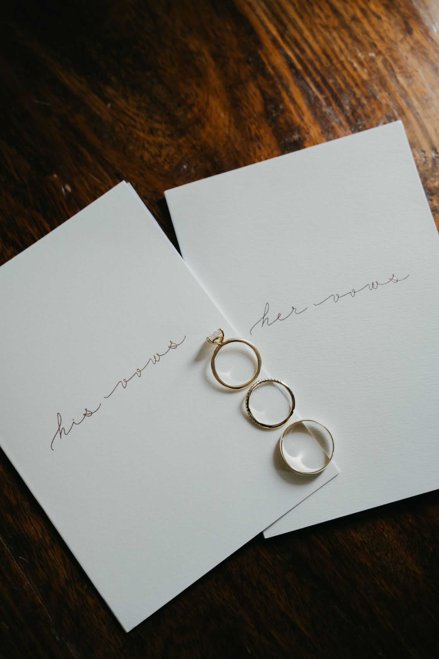 Wedding rings resting on his vows and her vows cards on a wooden table.