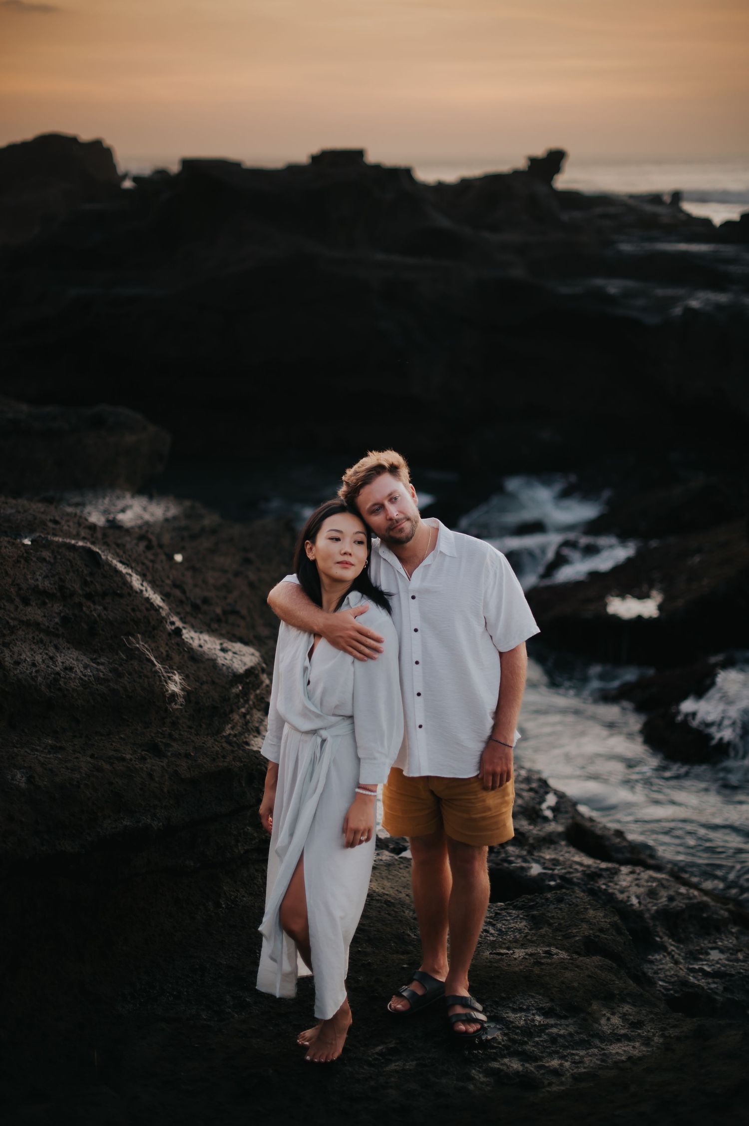Couple in white and brown outfits embrace on rocky coastline during sunset photoshoot.