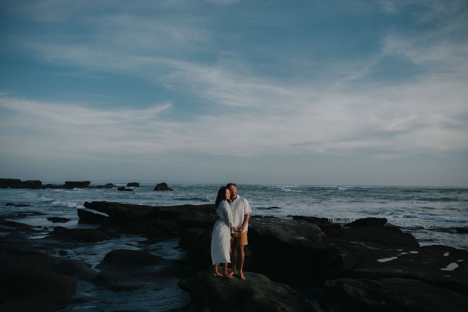 Romantic couple stands on rocky coastal cliffs at sunset with dramatic ocean waves crashing in the background.