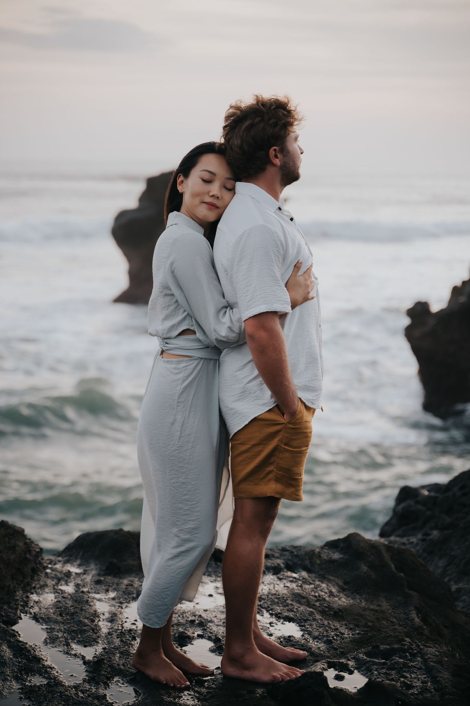 A couple embraces on rocky coastal cliffs at sunset as ocean waves crash against the shore.
