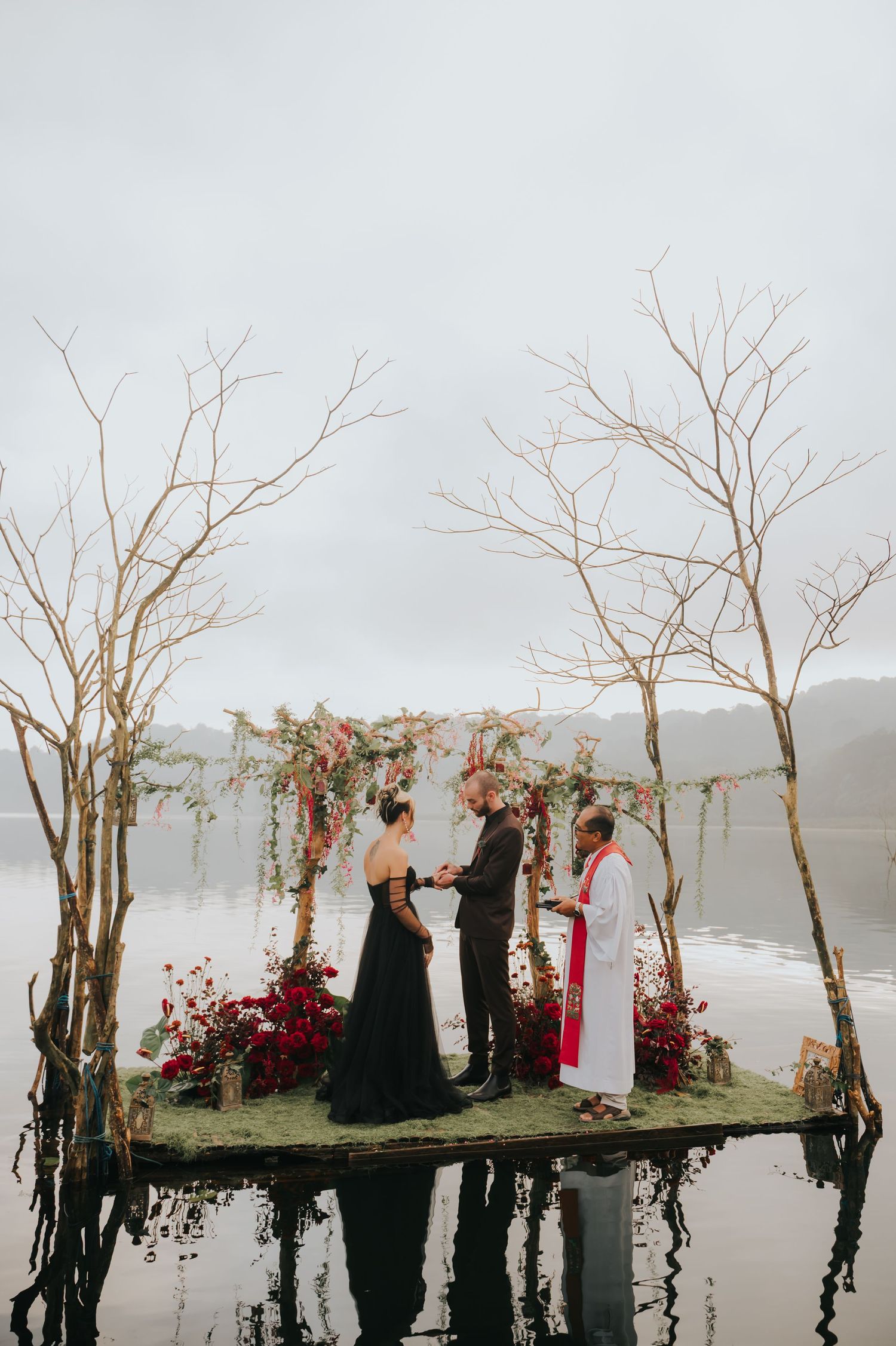 Moody wedding ceremony setup with bare trees and red floral arrangements reflected in still water.