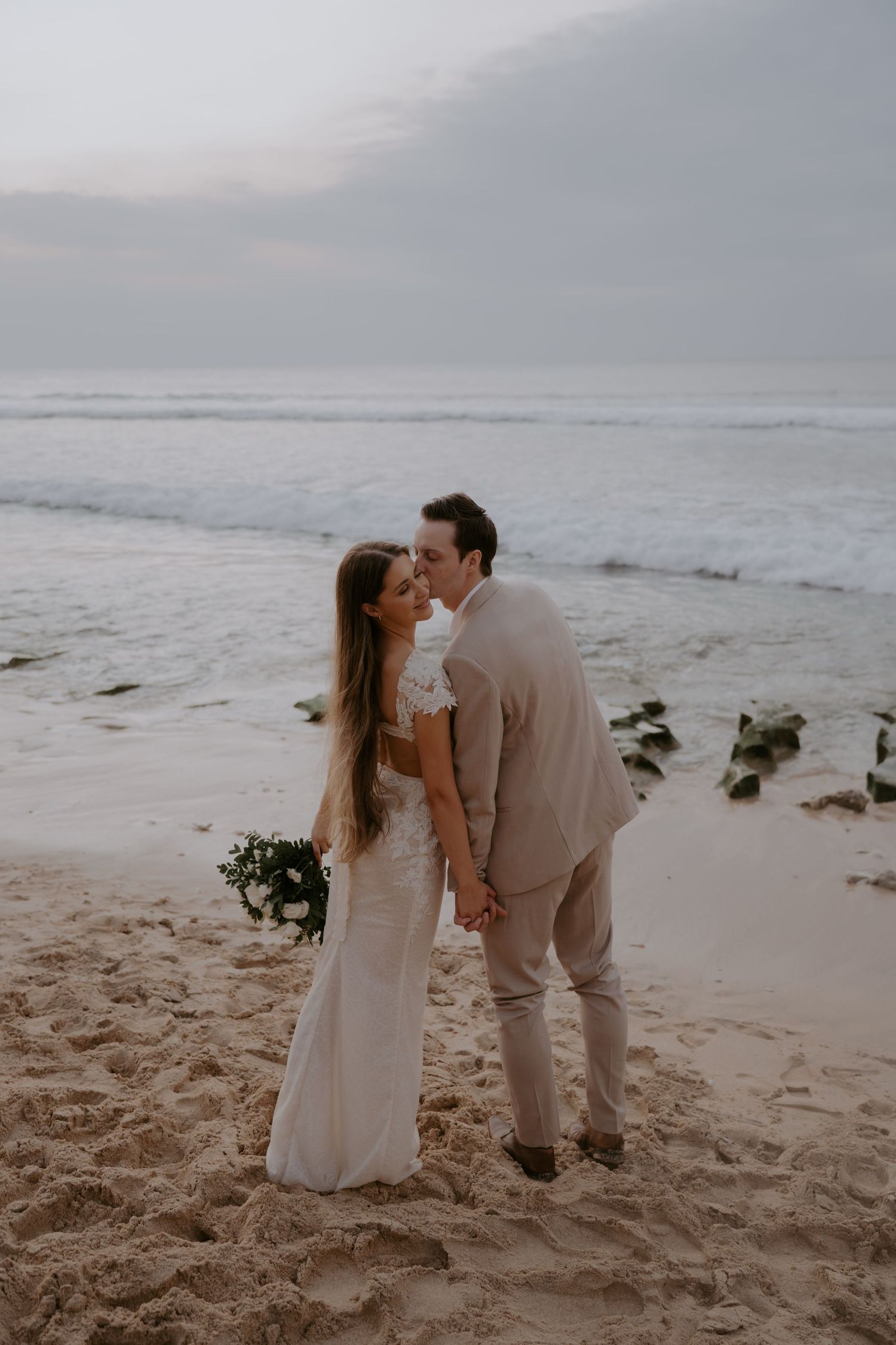 A romantic couple shares a tender moment on a misty beach at sunset, with waves crashing in the background.