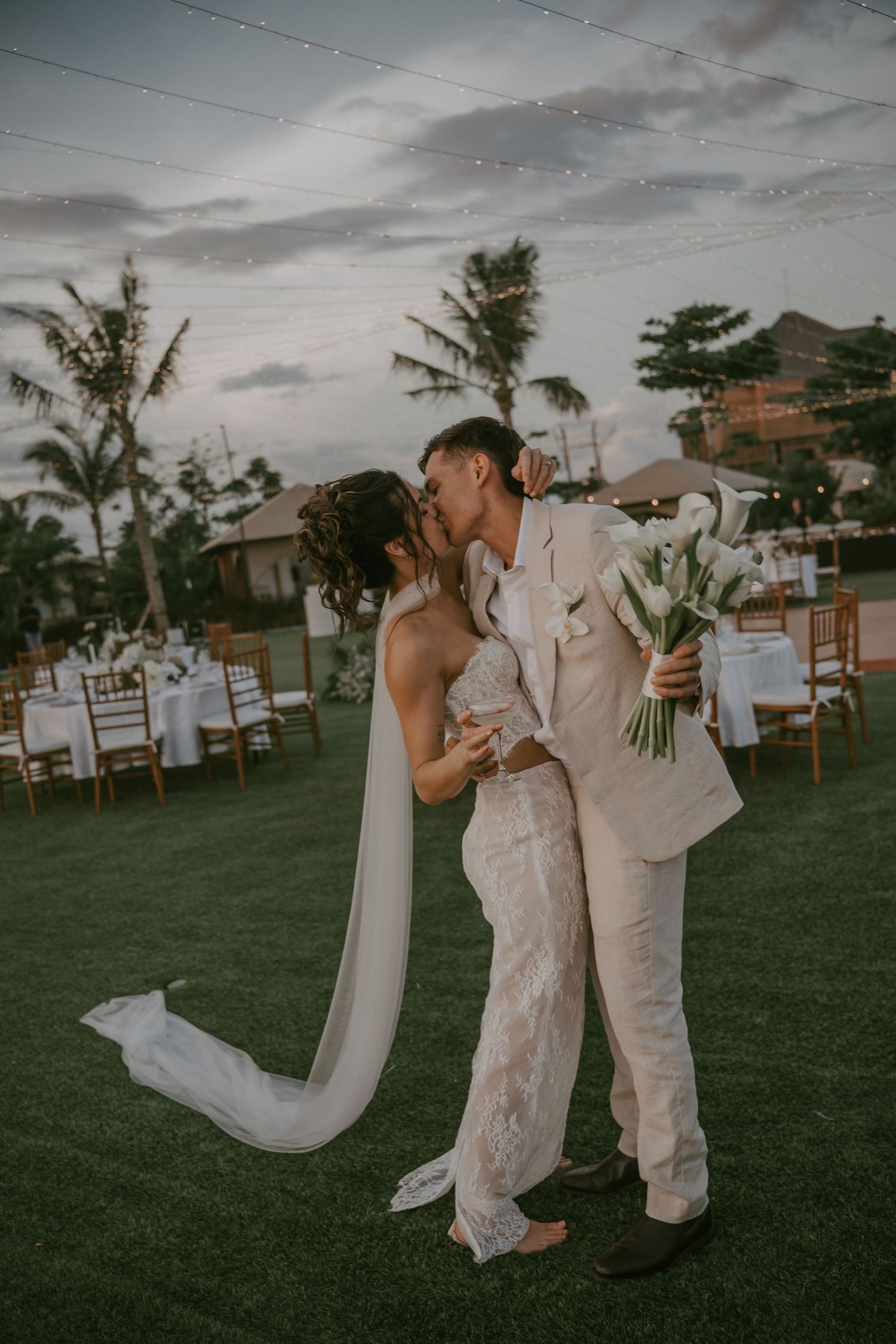 A couple shares a romantic moment at a sunset outdoor wedding reception with palm trees and white chairs in the background.