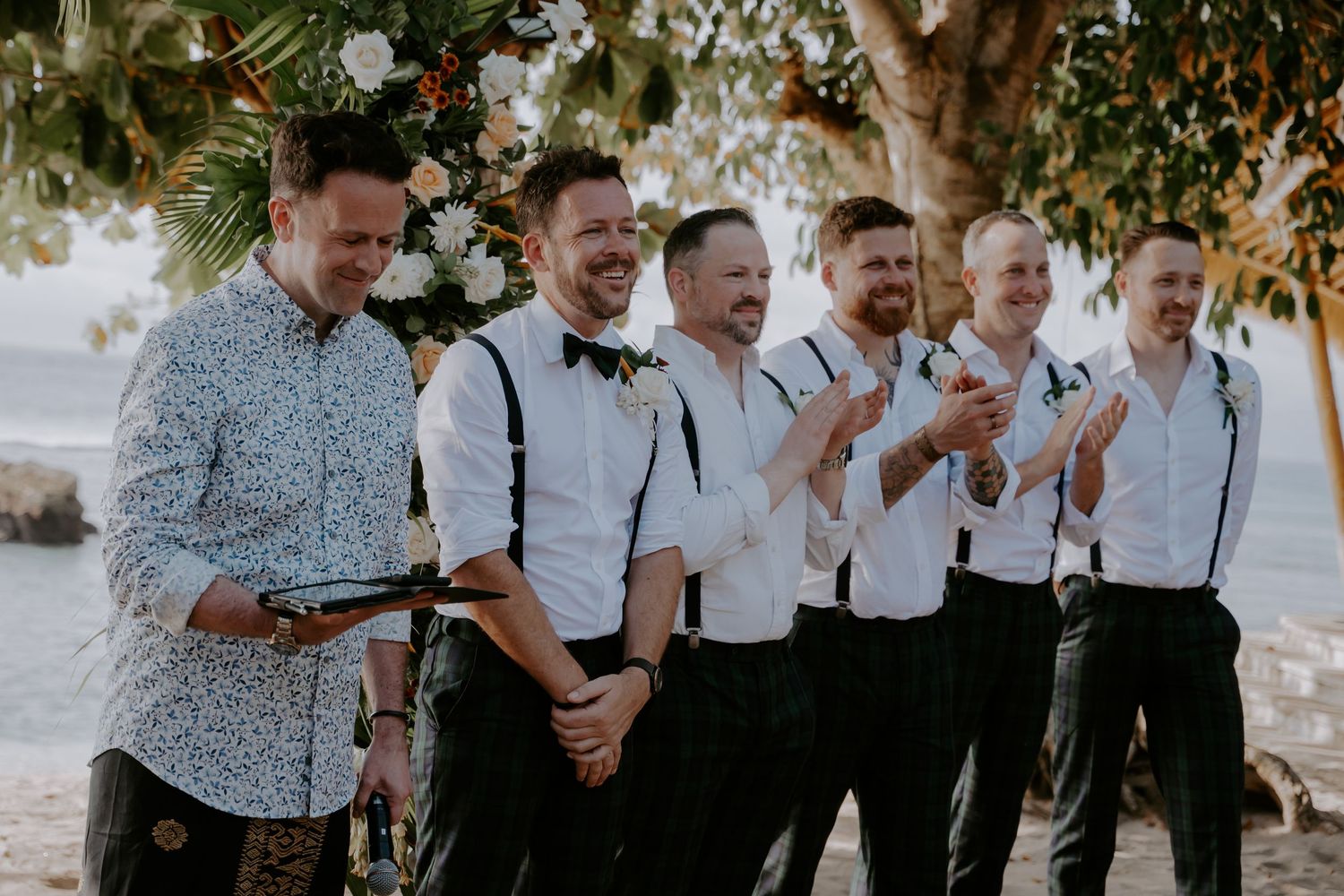Groomsmen in white shirts and black pants laughing together in a candid moment outdoors.