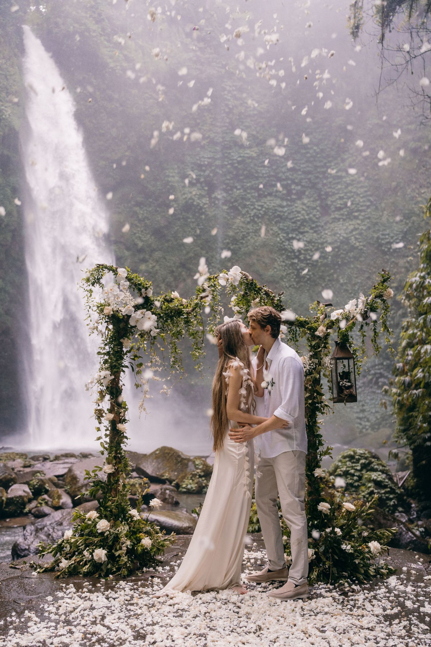 A romantic wedding ceremony takes place under a floral arch beside a majestic waterfall with flower petals falling in the air.